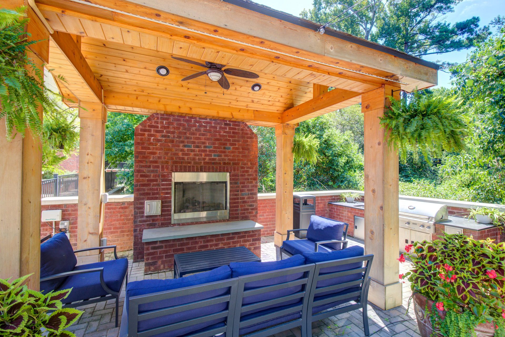 Patio with fireplace, seating, and wooden pergola. Blue furniture, brick and stone surfaces, greenery.