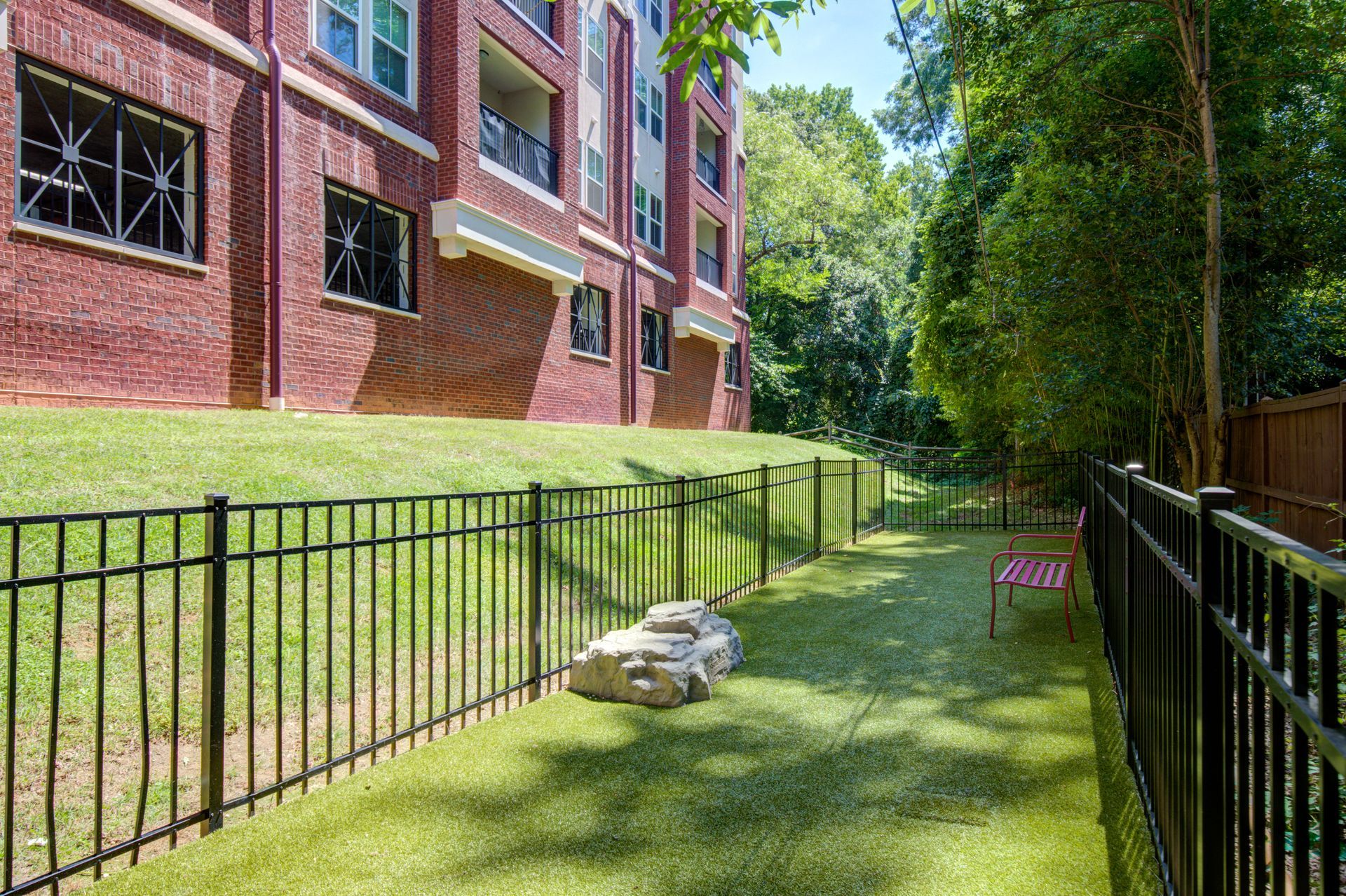 Dog park with black fence, green turf, brick building in background, and trees.