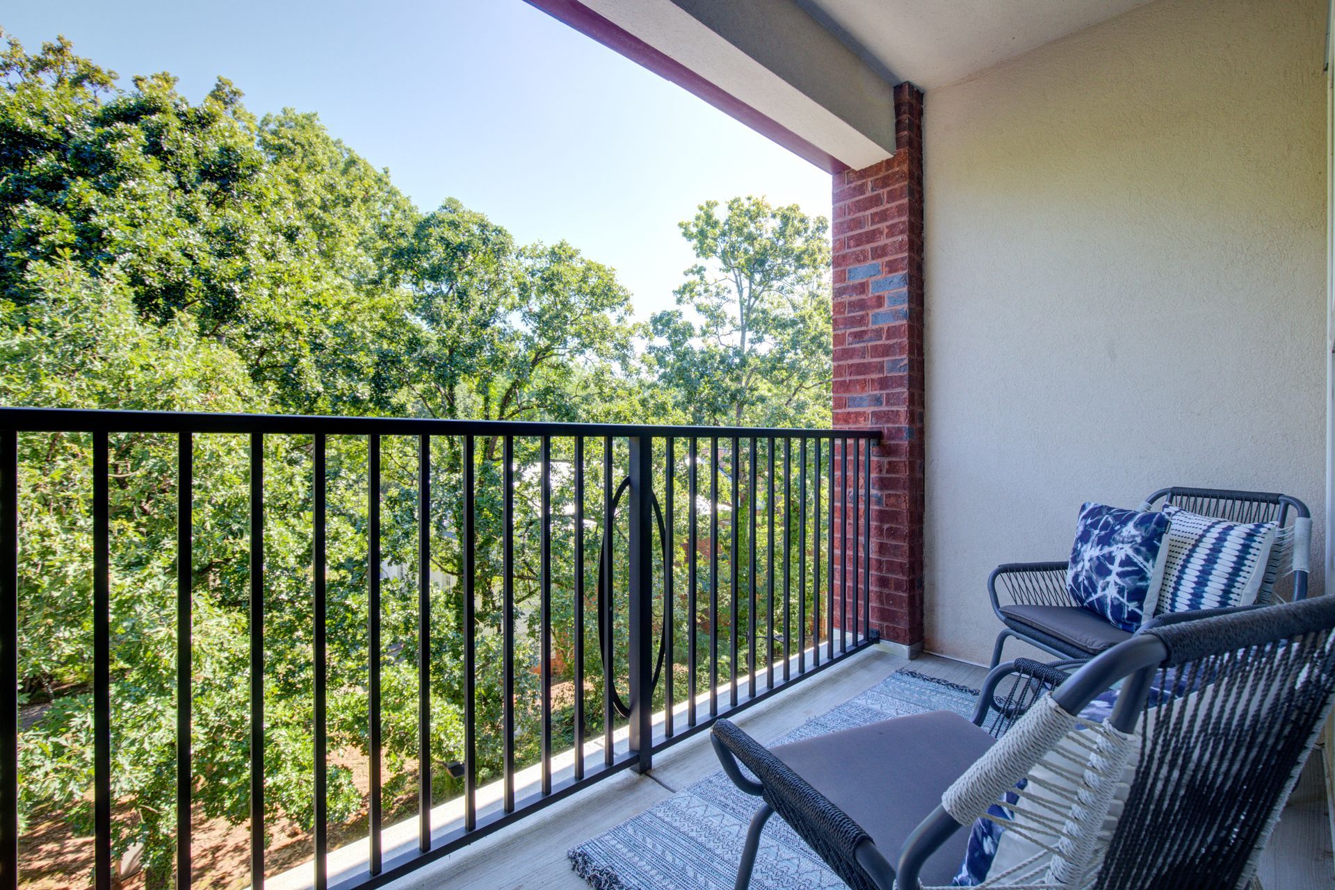 Balcony with black railing, two wicker chairs with blue pillows, overlooking green trees.