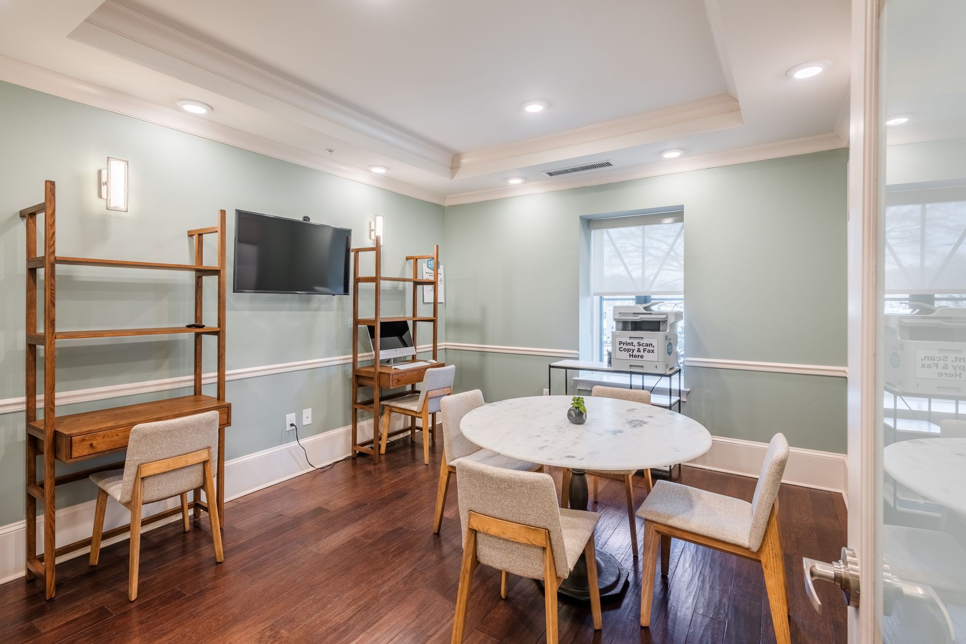 A light-filled room with round table and chairs, desks, a TV, and shelves.