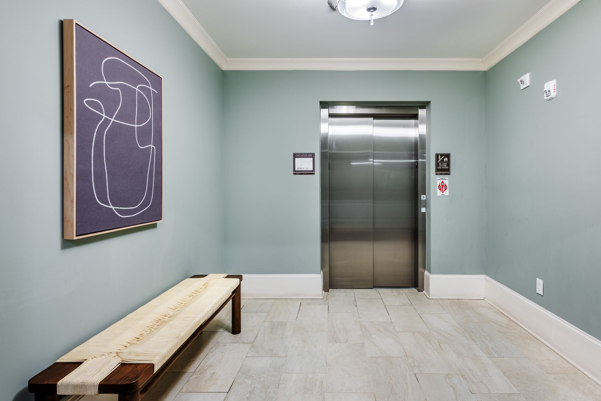 Hallway with elevator, bench, and artwork; blue-green walls, white trim, light floor.