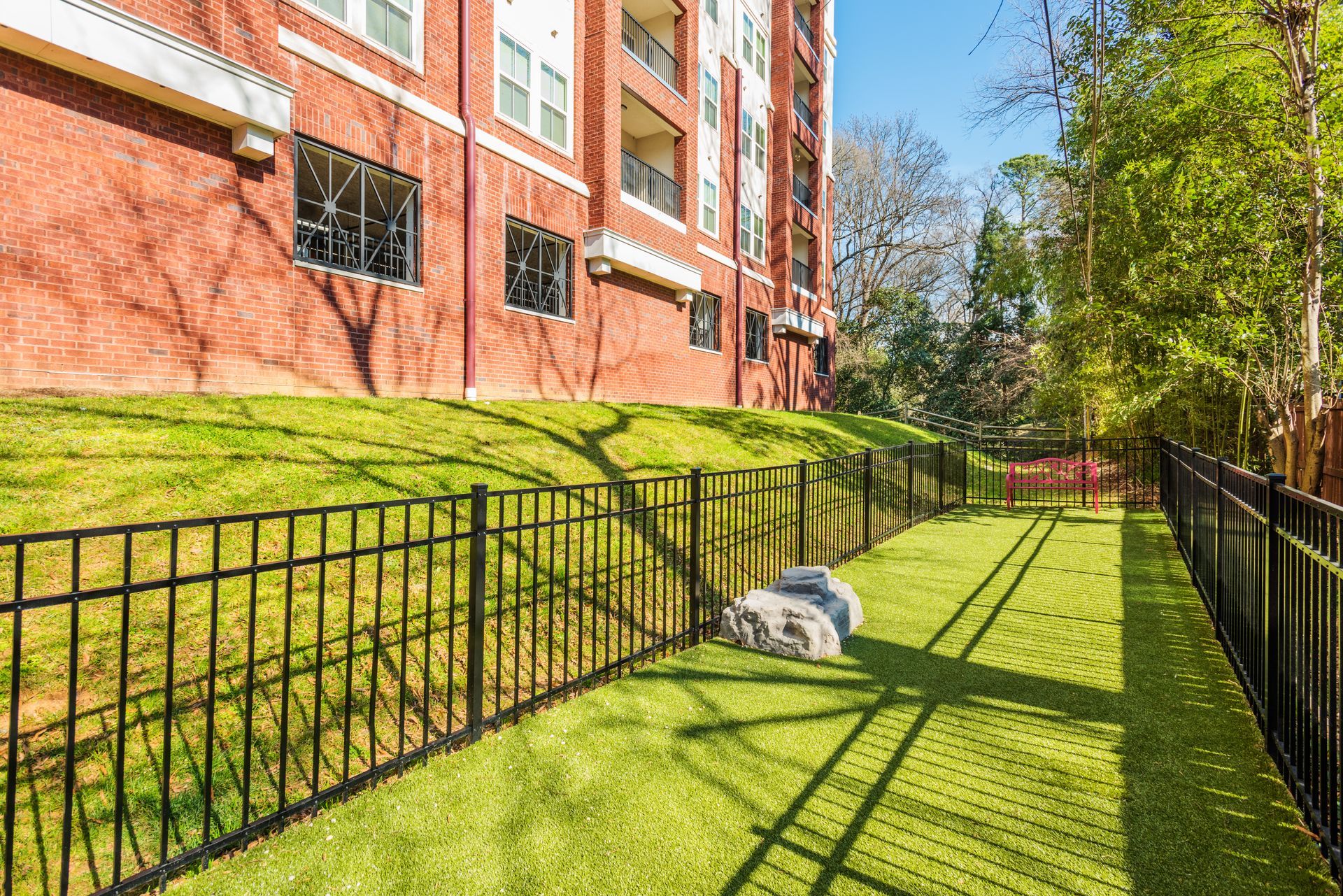 Dog park with black metal fence, green grass, and large stone. Apartment building in the background.