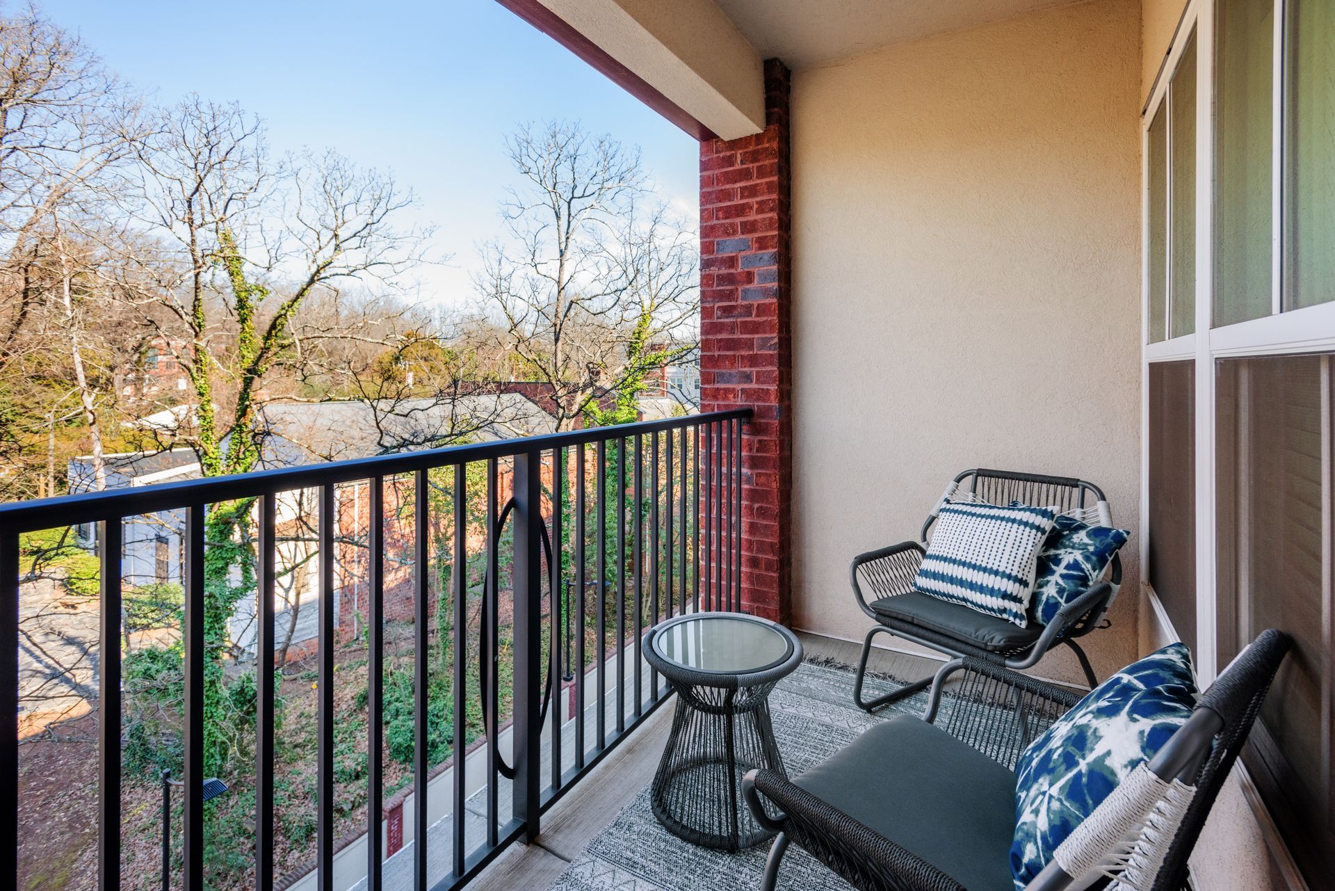 Balcony with two wicker chairs, small table, and decorative pillows overlooking a tree-lined street.