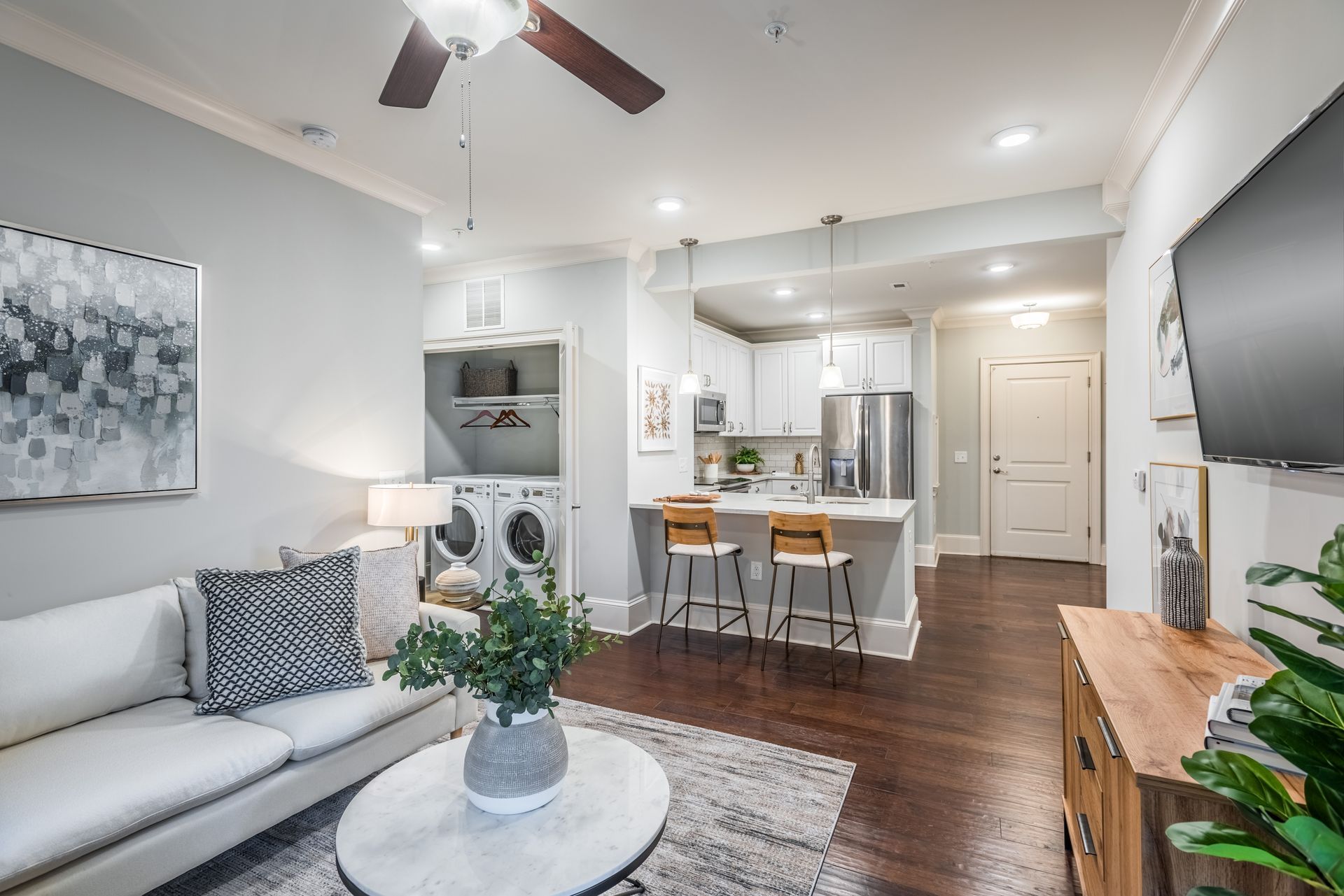 Open-concept apartment with living room, kitchen, and laundry. White walls, dark wood floors, light gray couch.
