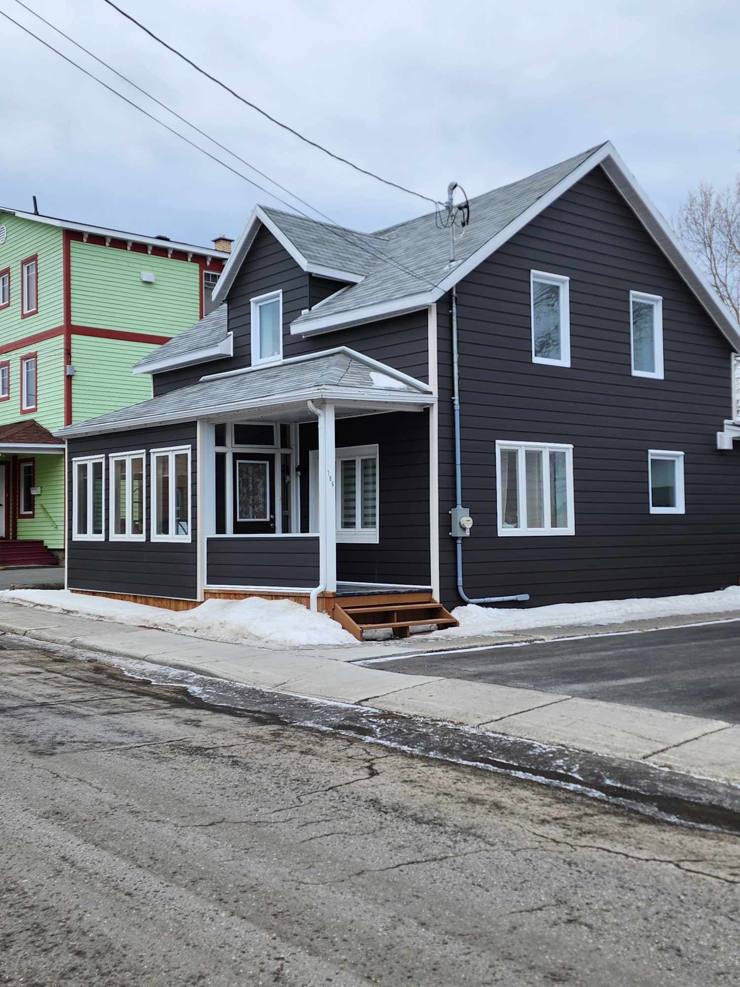 Une maison noire avec un porche se trouve au coin d'une rue à côté d'un bâtiment vert.