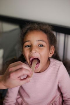 A little girl is being examined by a doctor with a wooden stick in her mouth.