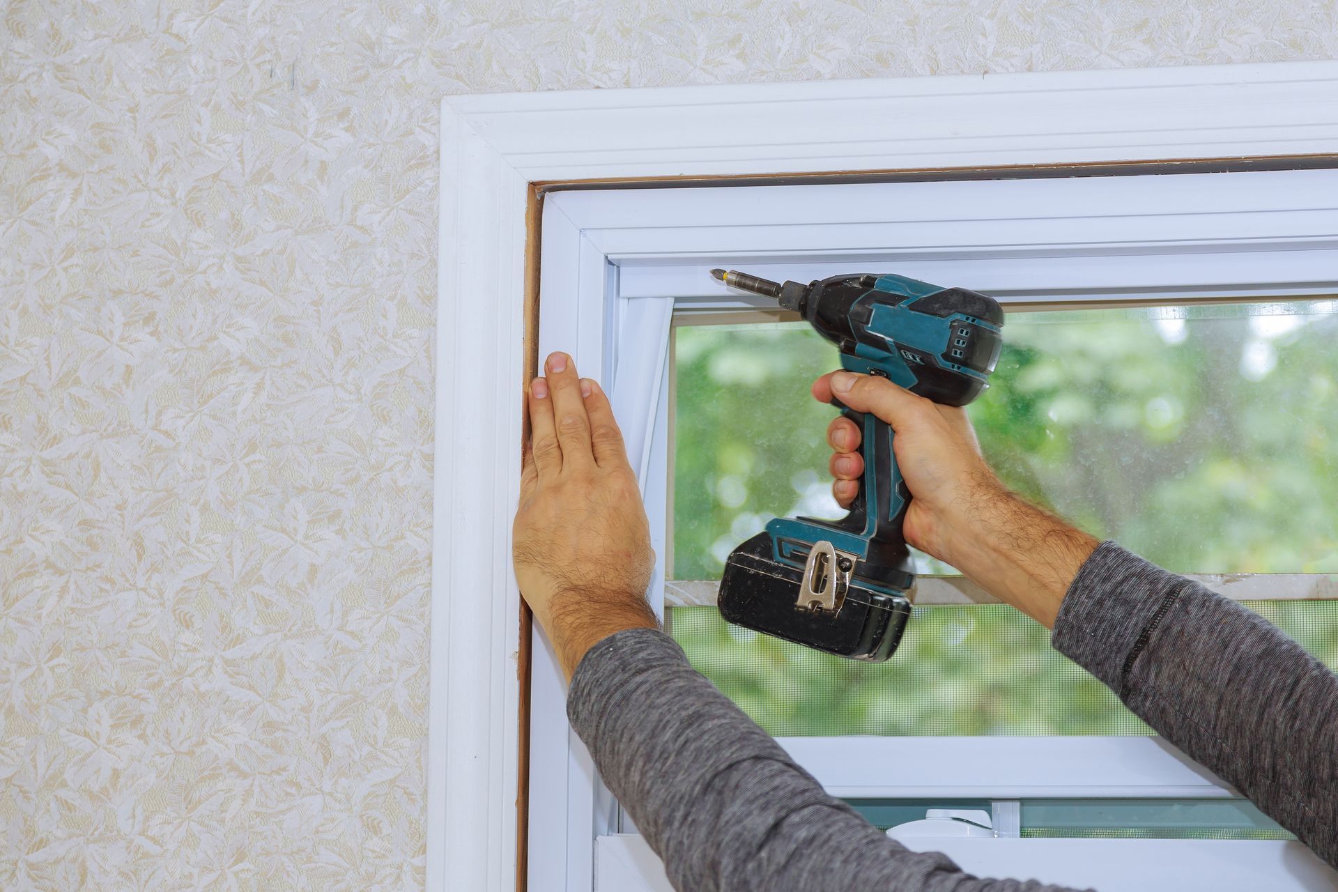 Technician using drill during residential glass installation on window frame inside home.