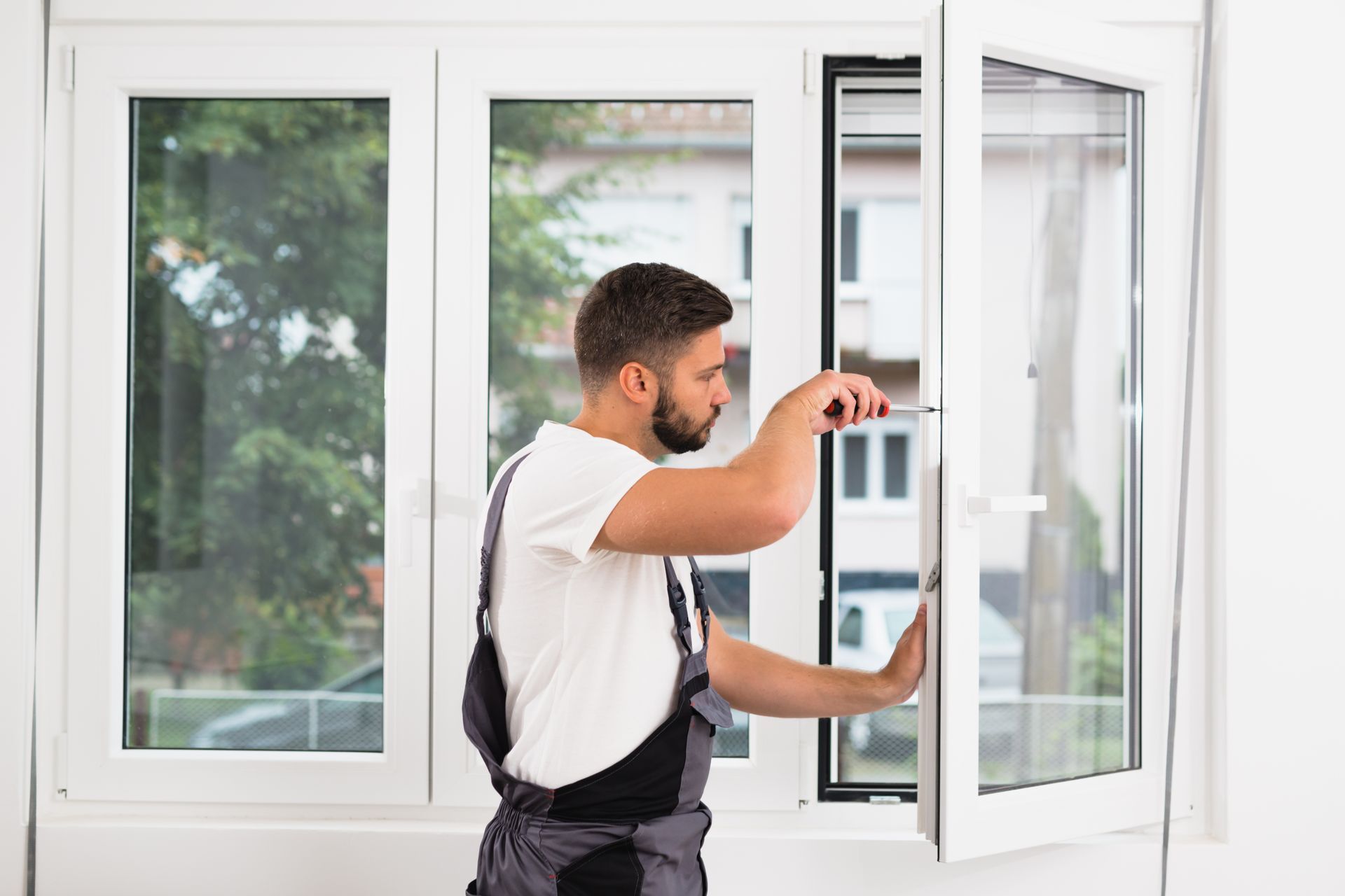 Worker adjusting a white-framed window with a screwdriver inside a bright room.