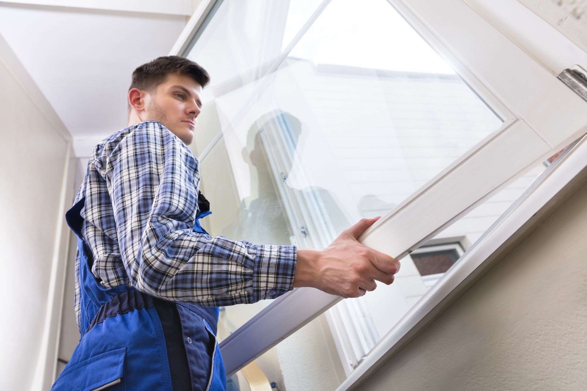 A man in blue overalls carries a white window, emphasizing window replacement.