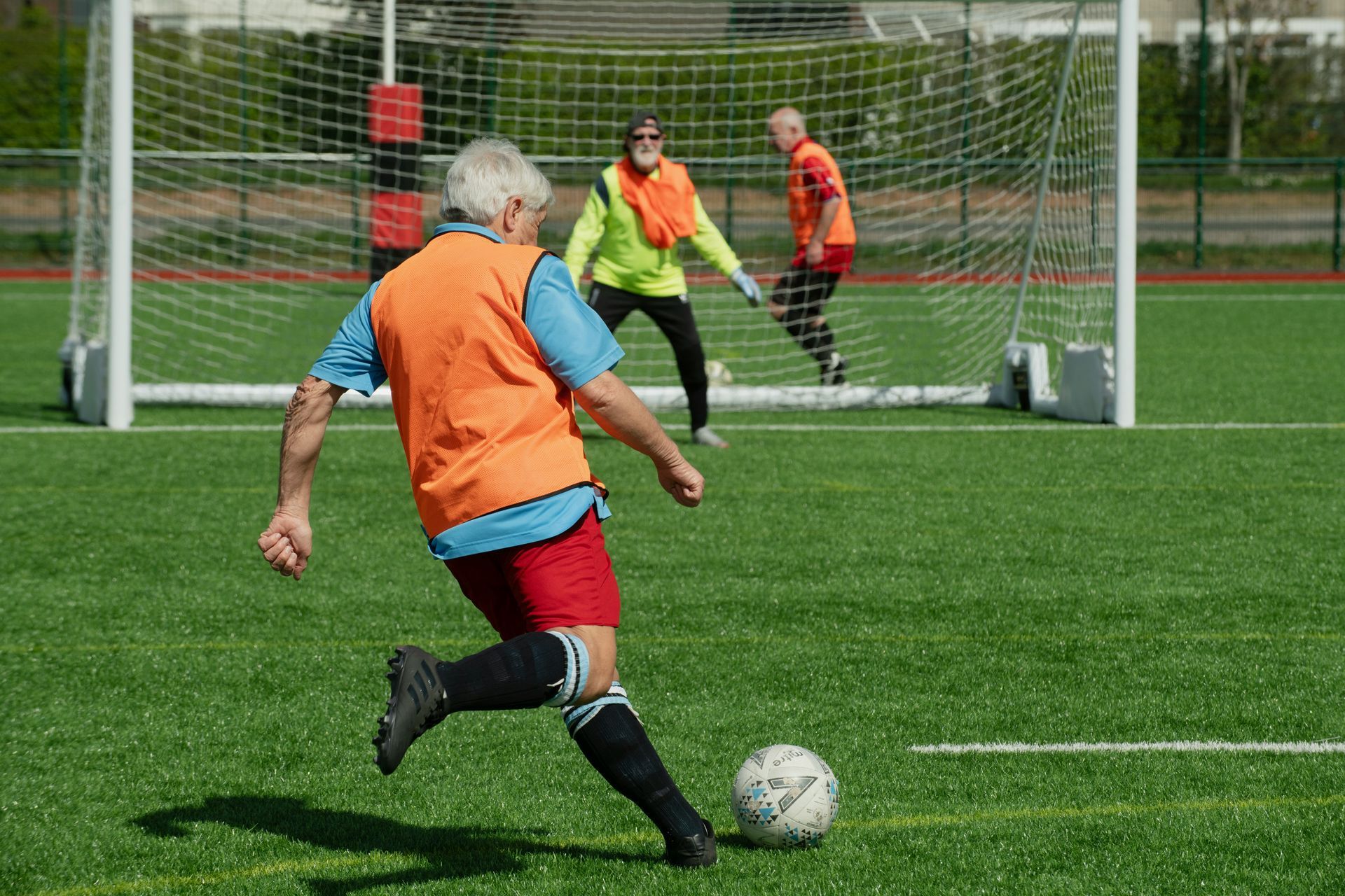Person in orange vest kicking soccer ball toward goal; two others in the background. Green field, sunny day.