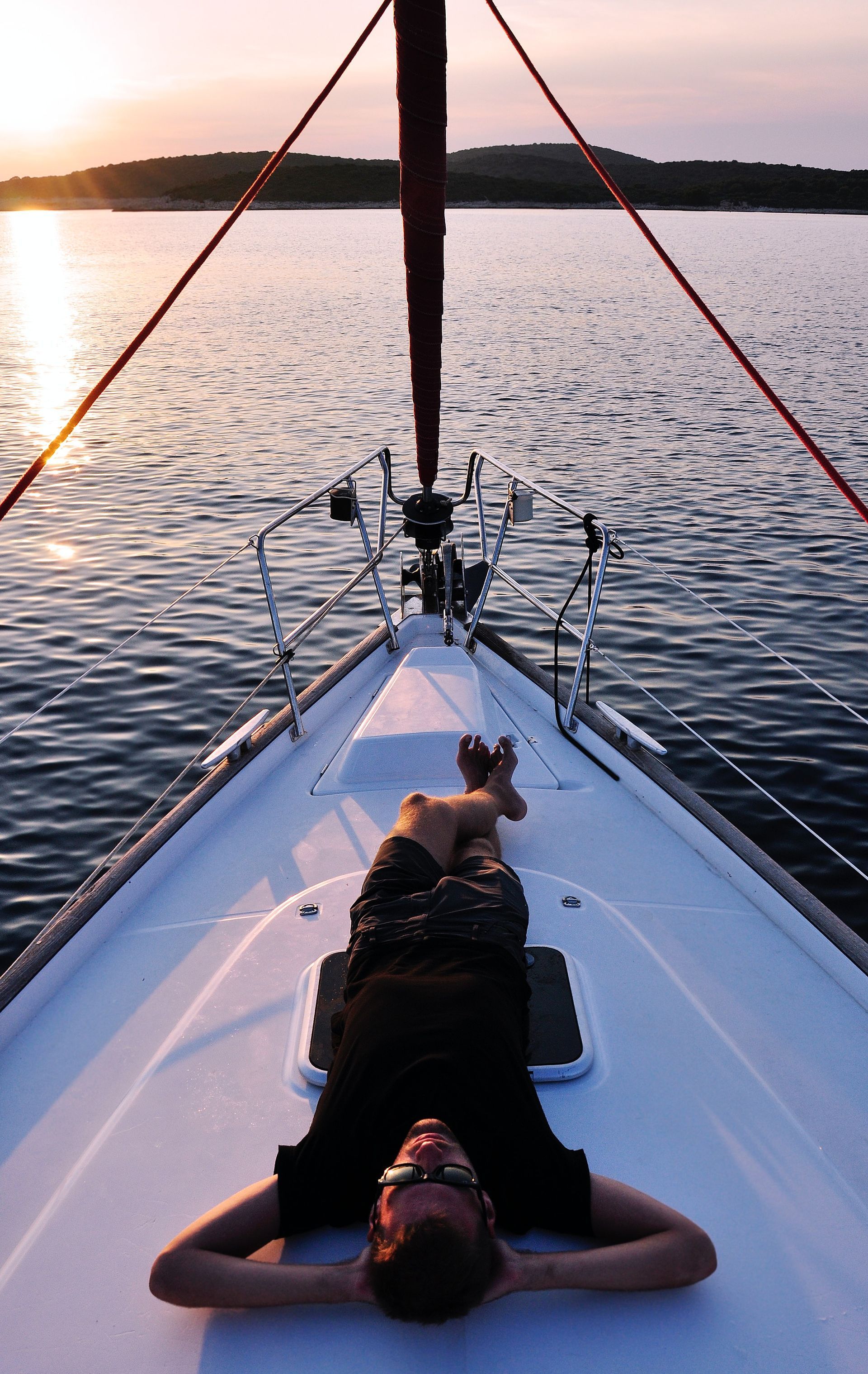 Man relaxing on a sailboat deck, looking up at sunset over calm water.