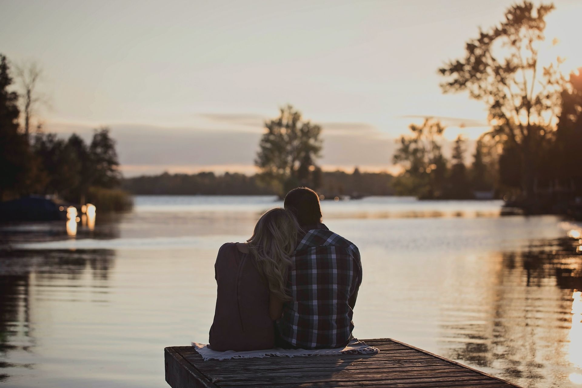 Couple sitting on a wooden dock, facing a lake at sunset.