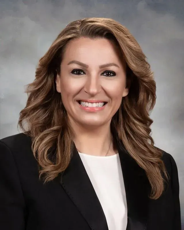 Woman with brown wavy hair smiling in a black blazer and white top.