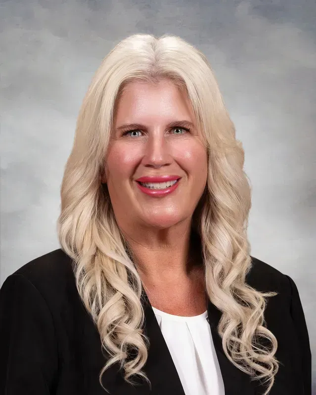 Woman with long, wavy blonde hair smiles, wearing a black blazer and white blouse, against a blurred background.