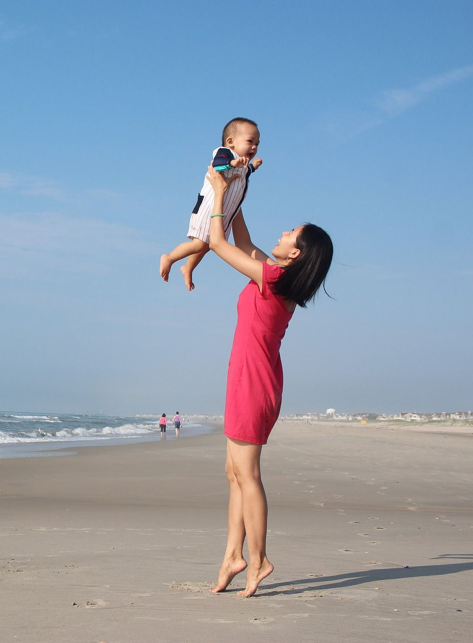 Woman in red dress holding a baby in the air on a beach; blue sky and ocean in background.