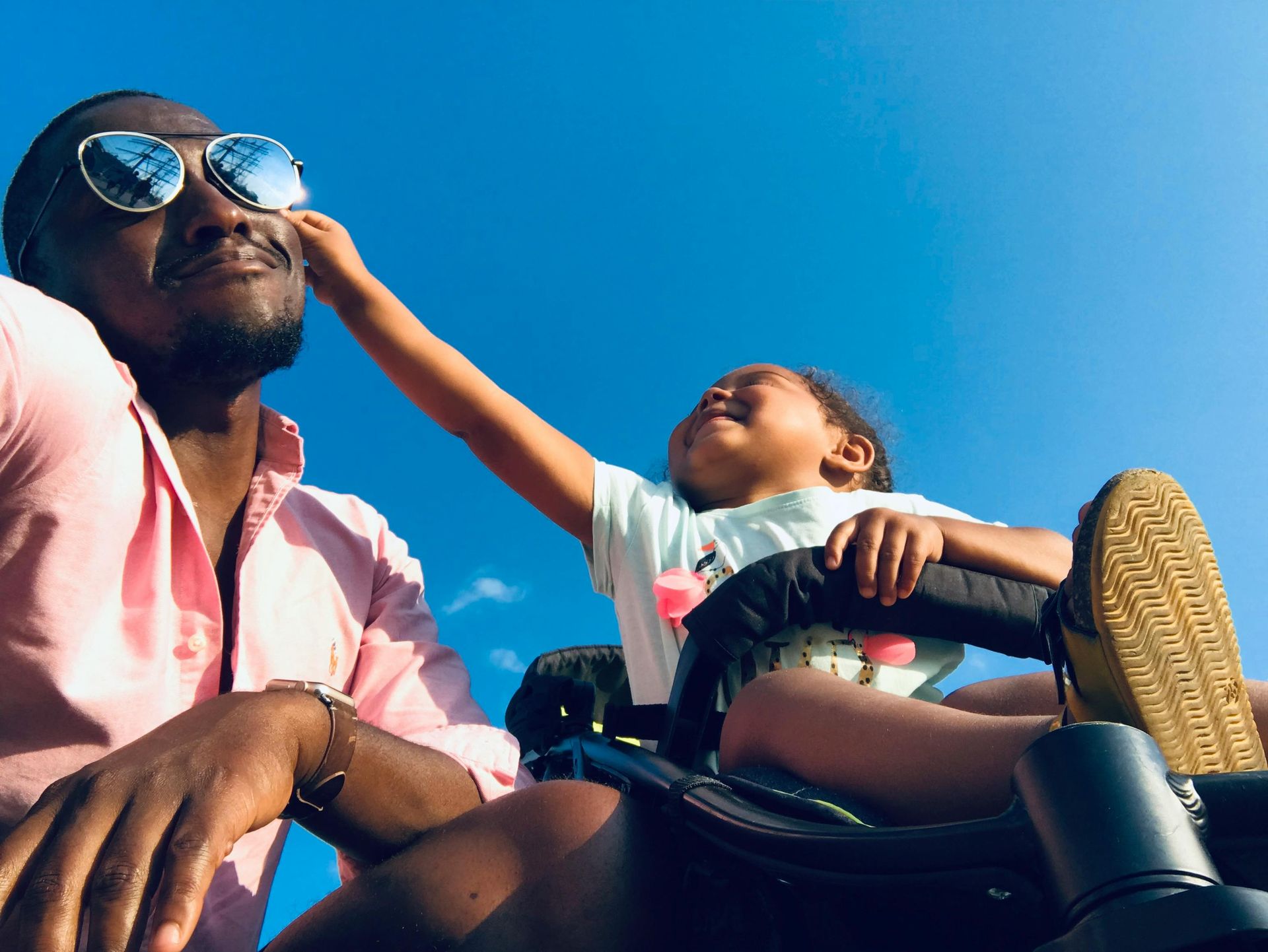 Man in sunglasses, seated, smiles as child touches his face, blue sky.