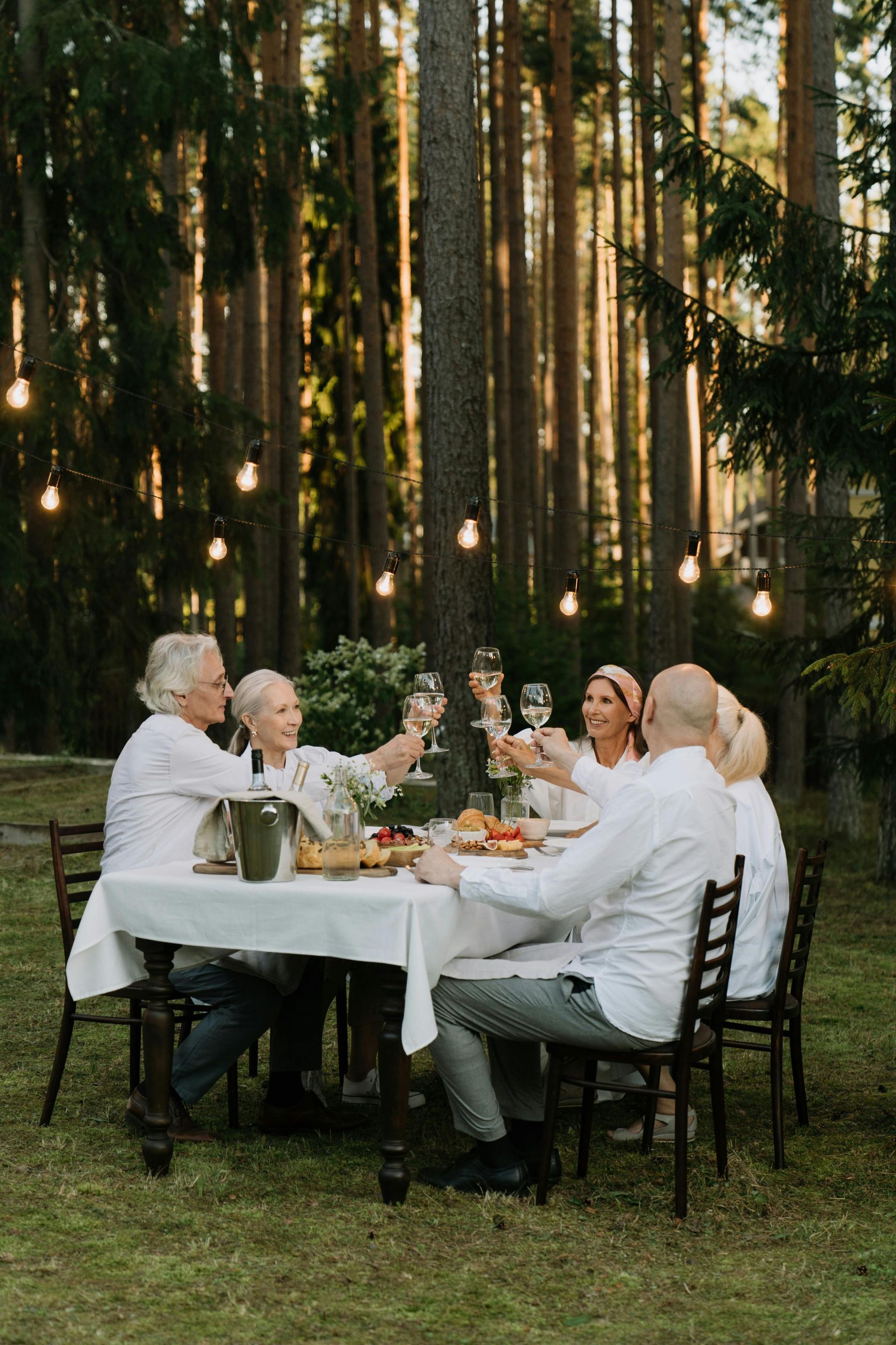 People toasting drinks at a table in a forest, lit by string lights.