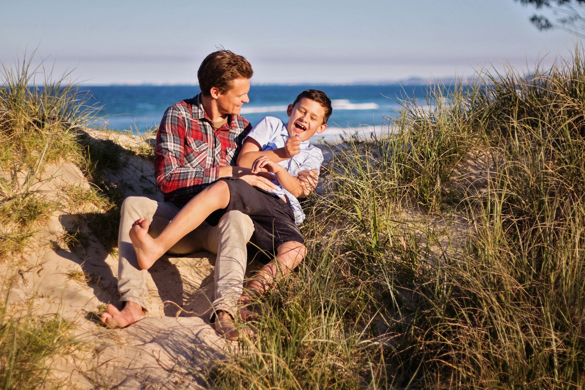 Two people laughing together on a sand dune, near the ocean. One is tickling the other.