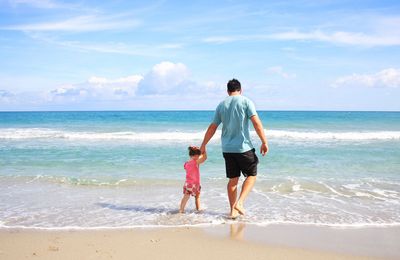 Man and child walk into the ocean, holding hands, on a sunny beach.