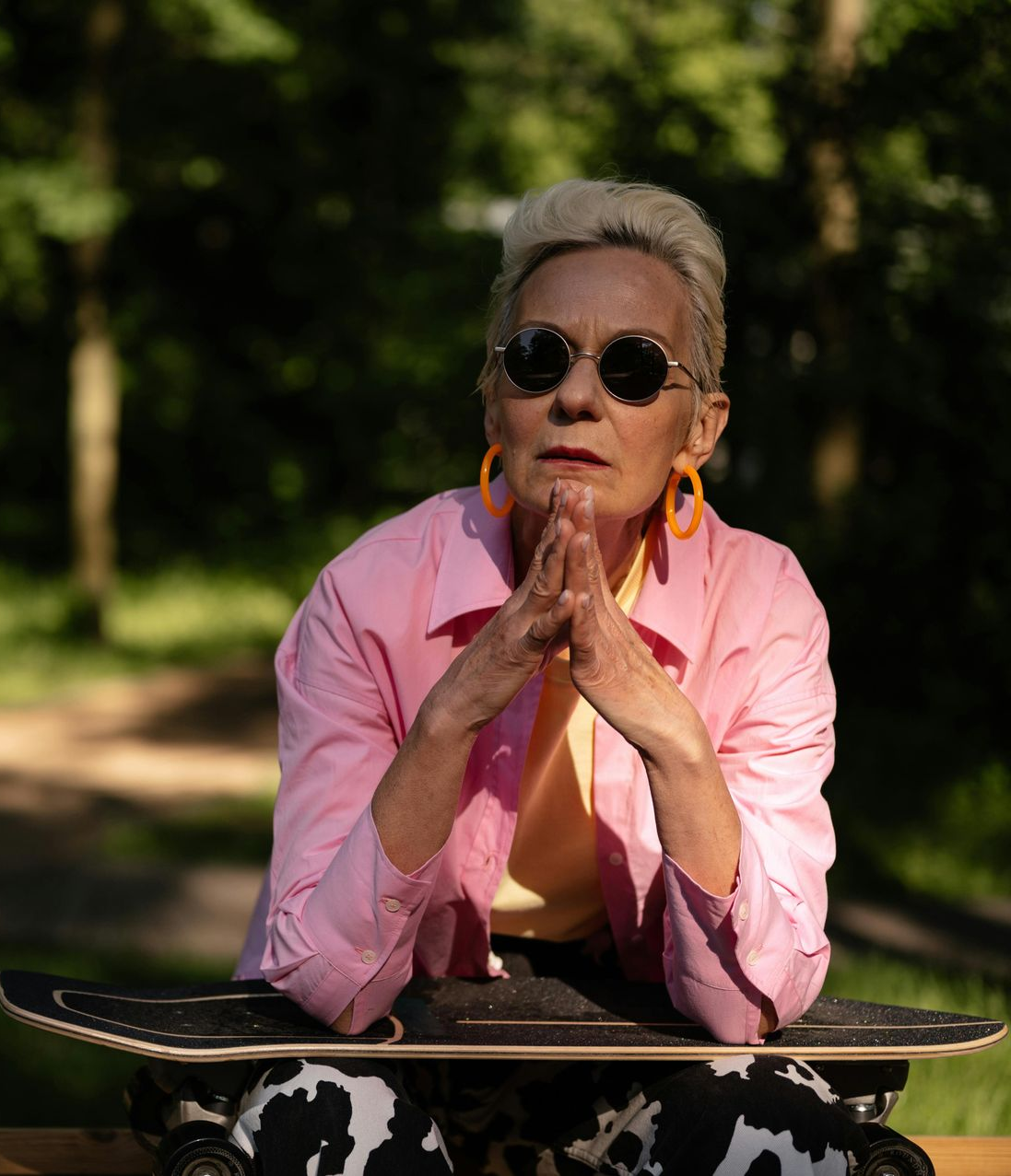 Person in pink jacket and sunglasses sits on skateboard, hands clasped, outdoors.