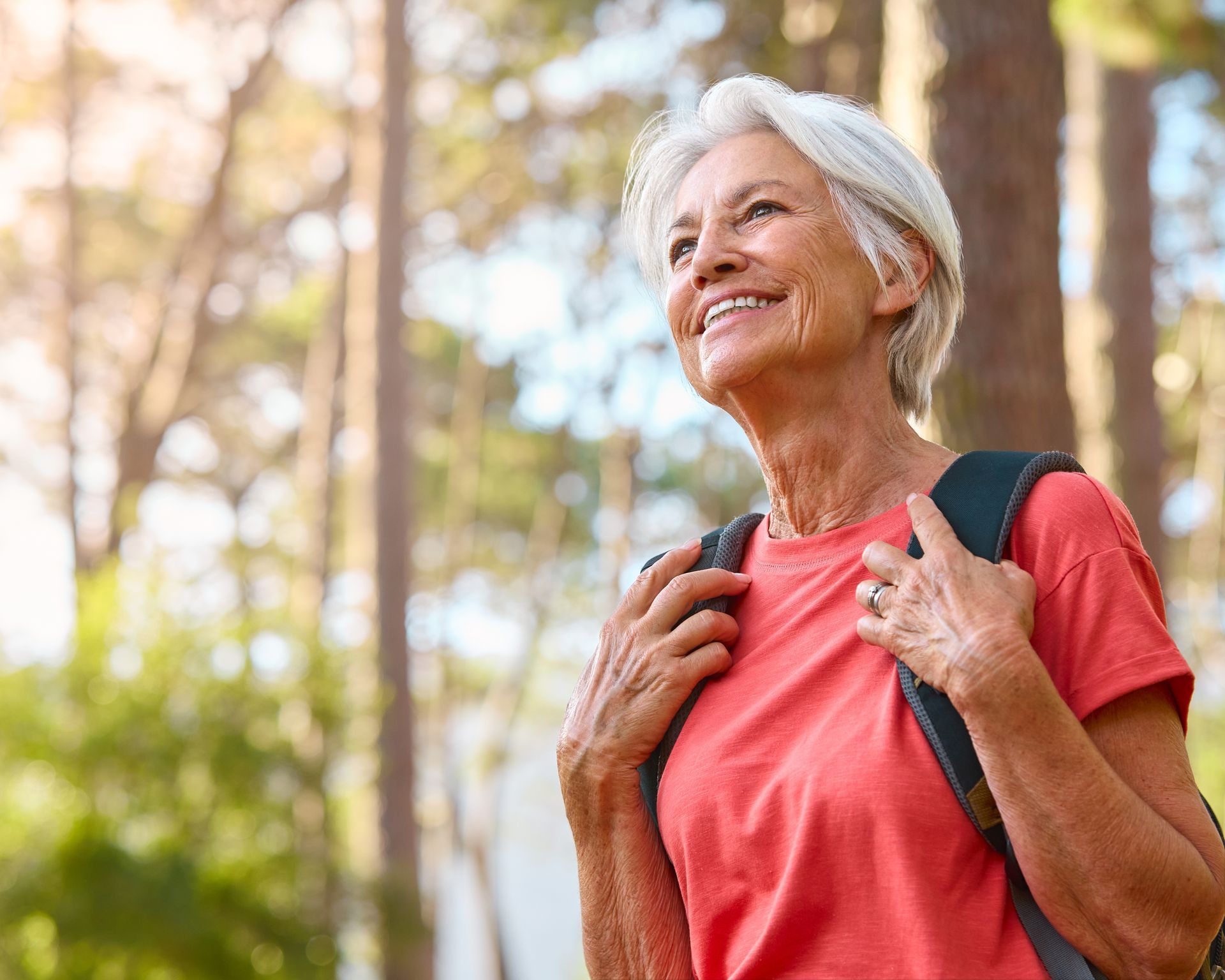Older person with gray hair wearing a red shirt and backpack smiles while hiking in a sunny forest.