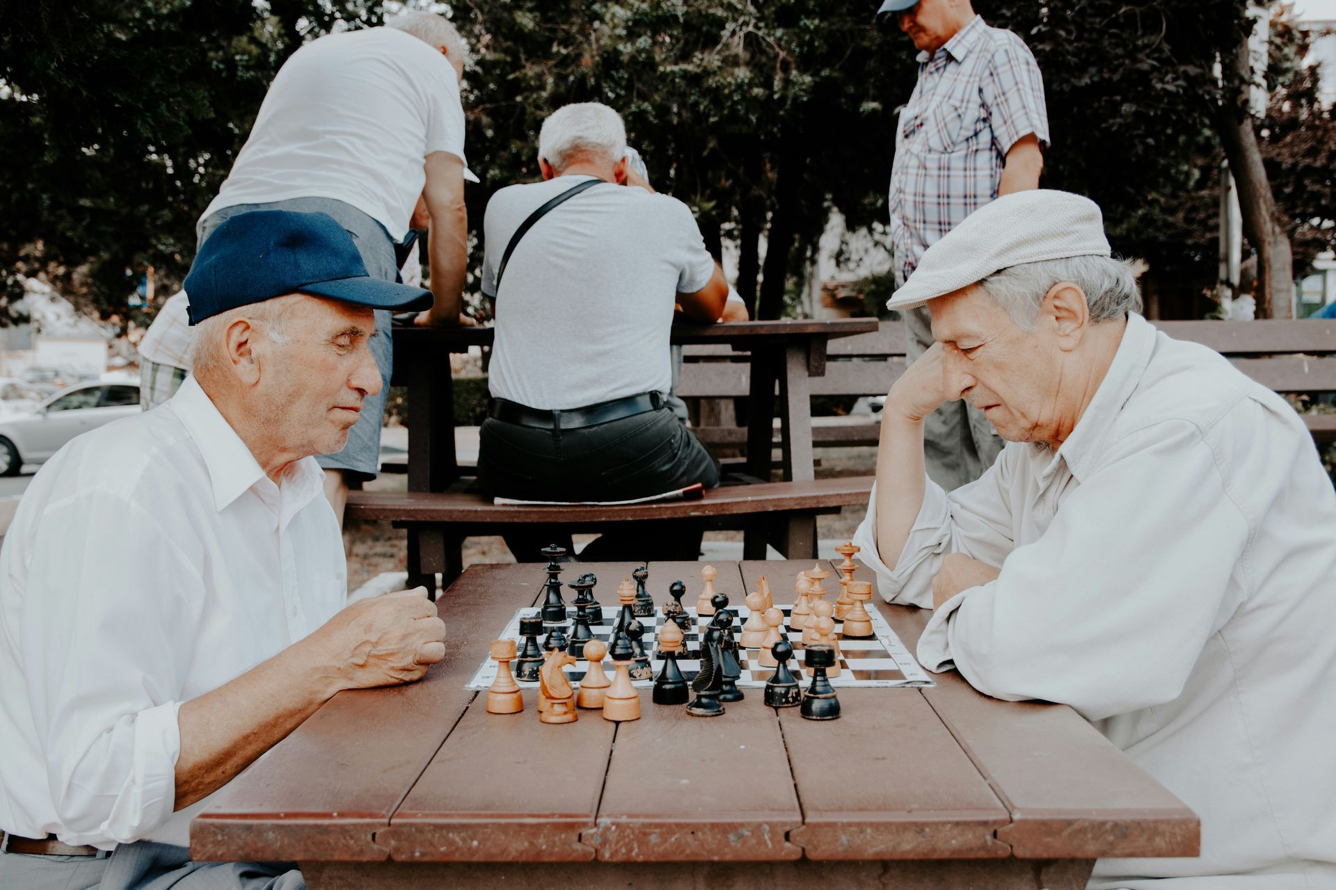 Two men playing chess at a park table, other people nearby.