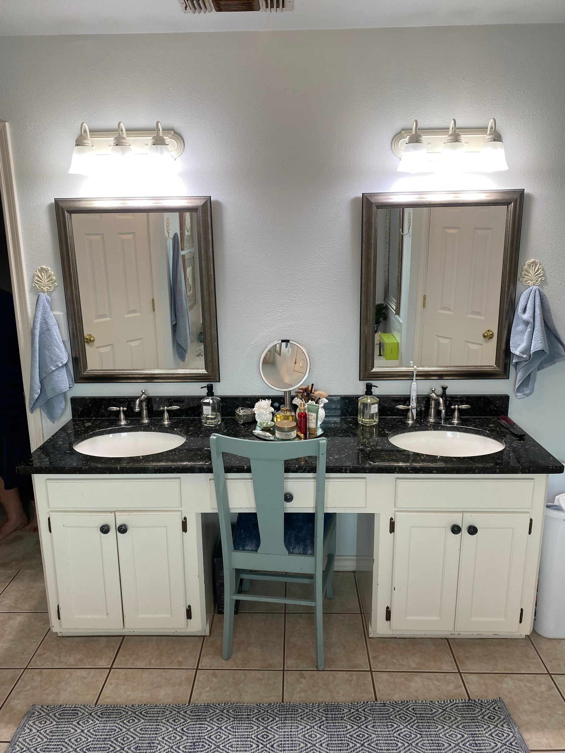 A bathroom with two sinks , two mirrors and a chair.