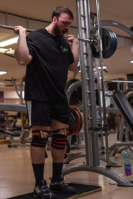 Nicola Voltolina in palestra, con indosso una canottiera nera, seduto con le mani giunte. Attrezzi da palestra visibili sullo sfondo.
