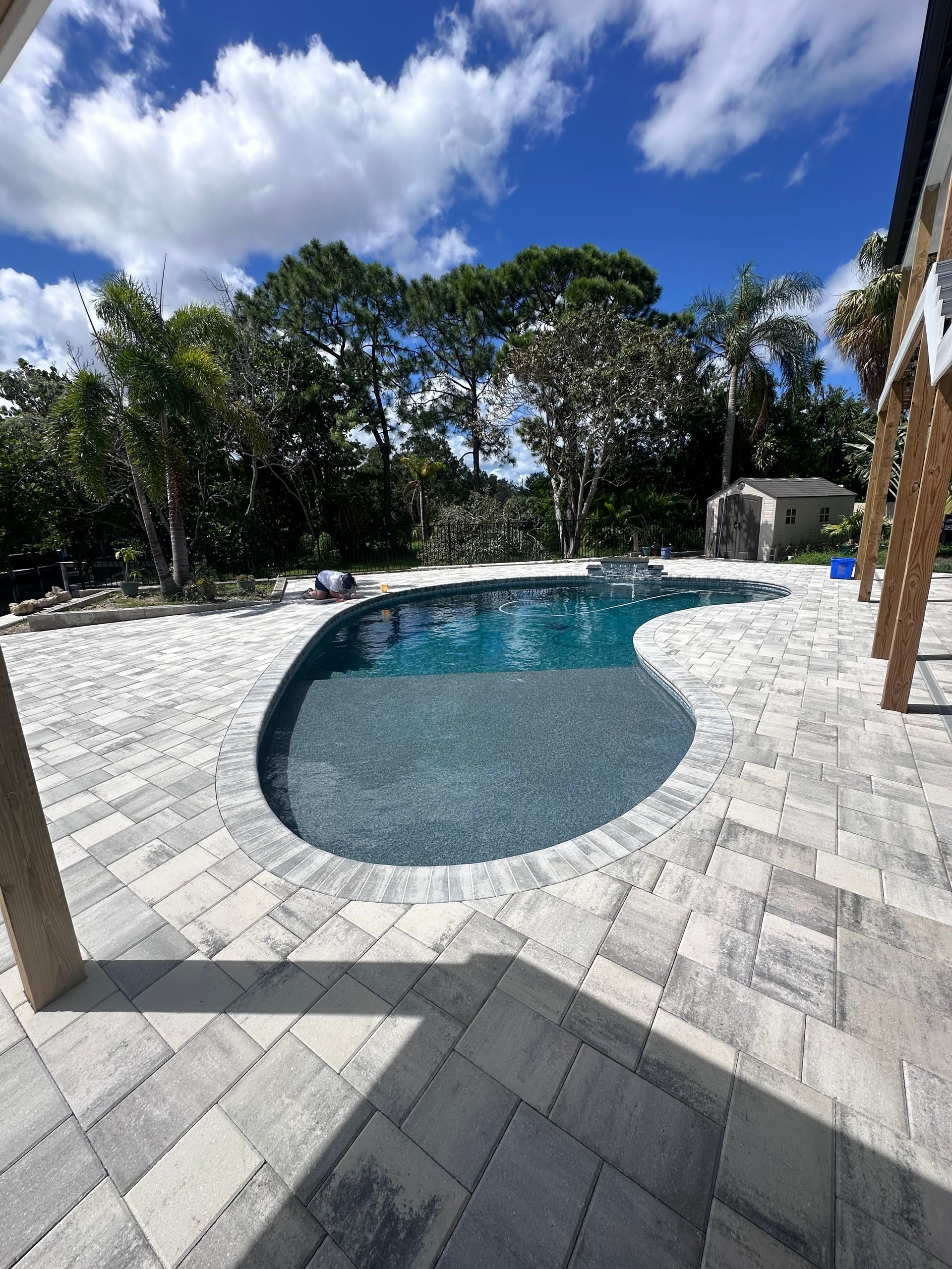 Pool with dark blue water surrounded by light gray paving stones. Trees and blue sky background.