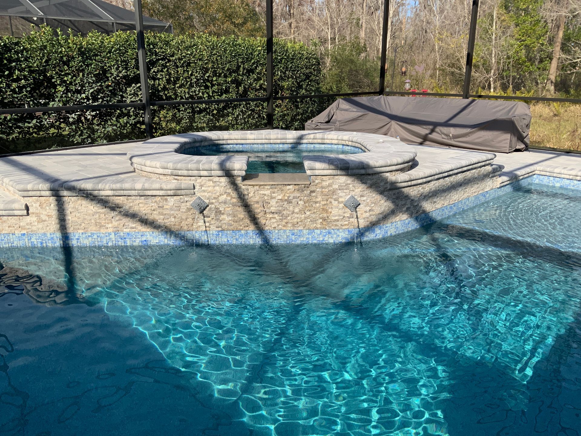 Pool with built-in hot tub, surrounded by stone and lush greenery, under a sunny sky.