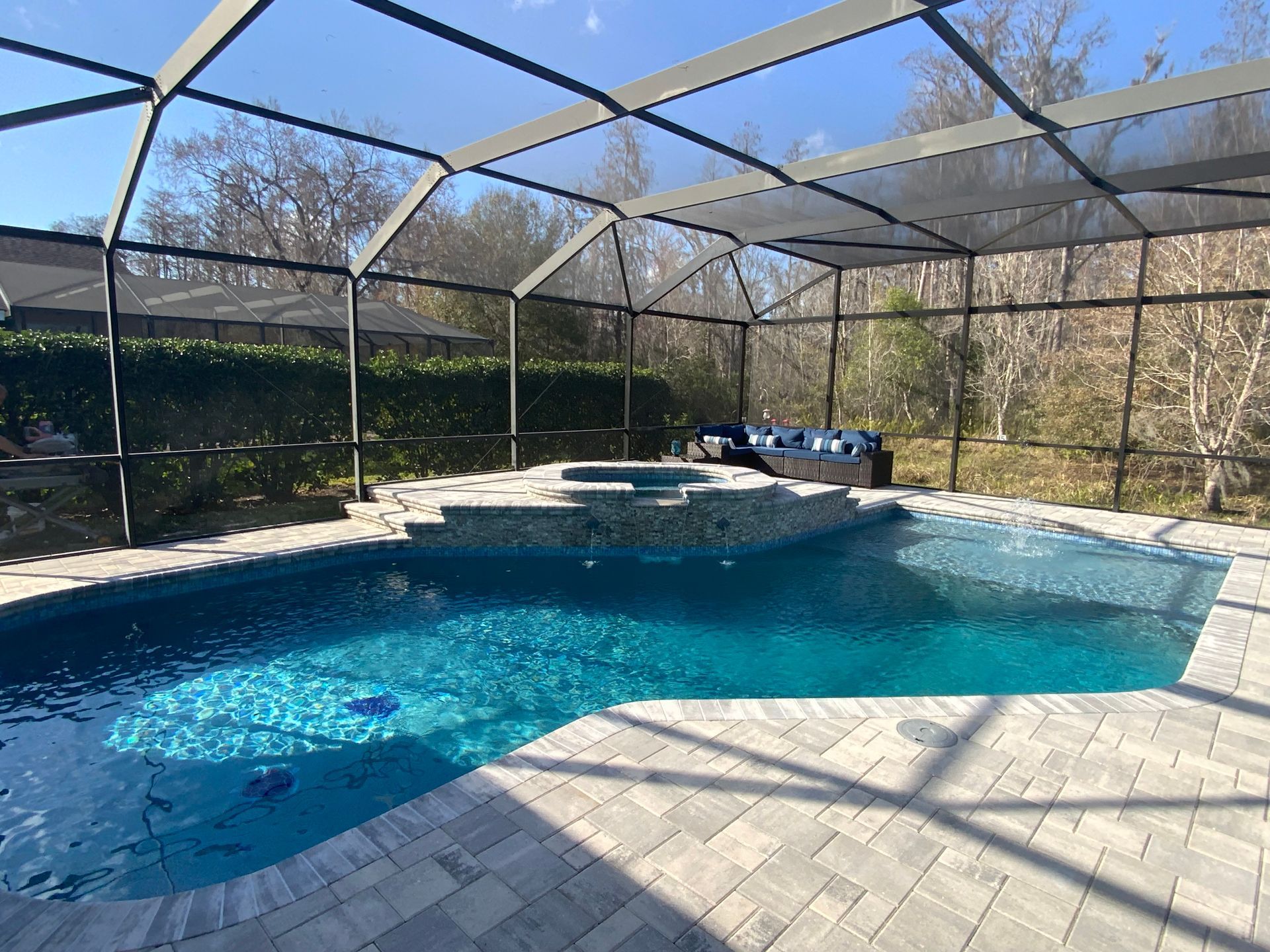 Swimming pool with a spa, screened enclosure, and stone patio. Blue water and a natural setting.
