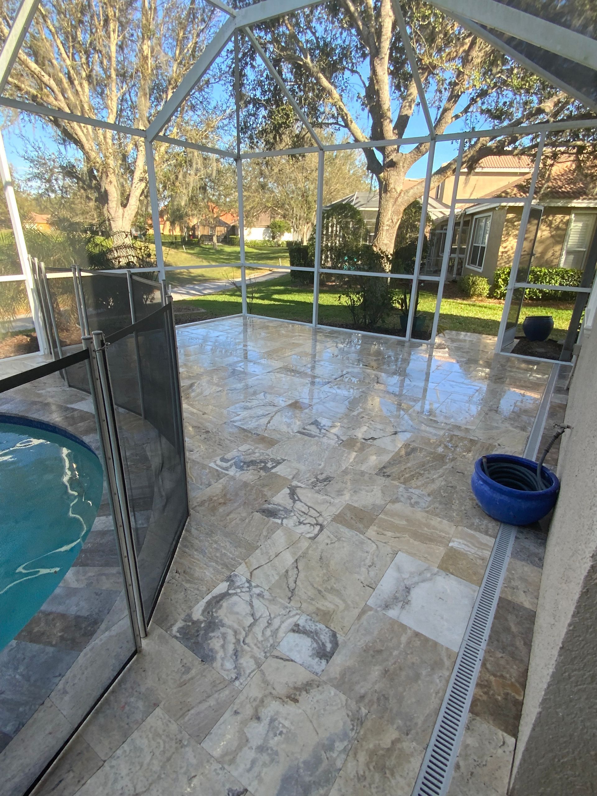 Tiled patio with a screened enclosure. A pool and a drain are visible. Trees and a sunny sky in the background.