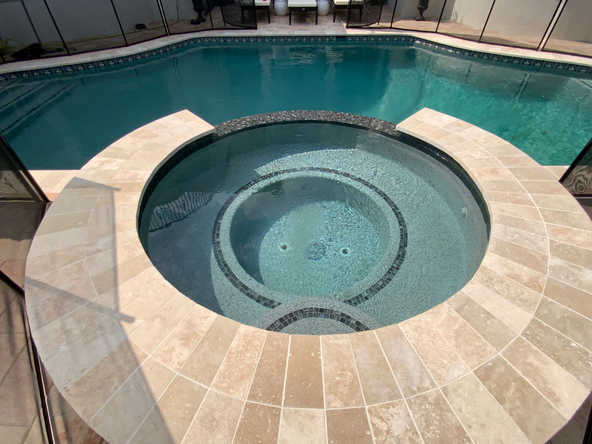 Pool with a round hot tub in the center, surrounded by tan tile. Clear blue water in the pool.