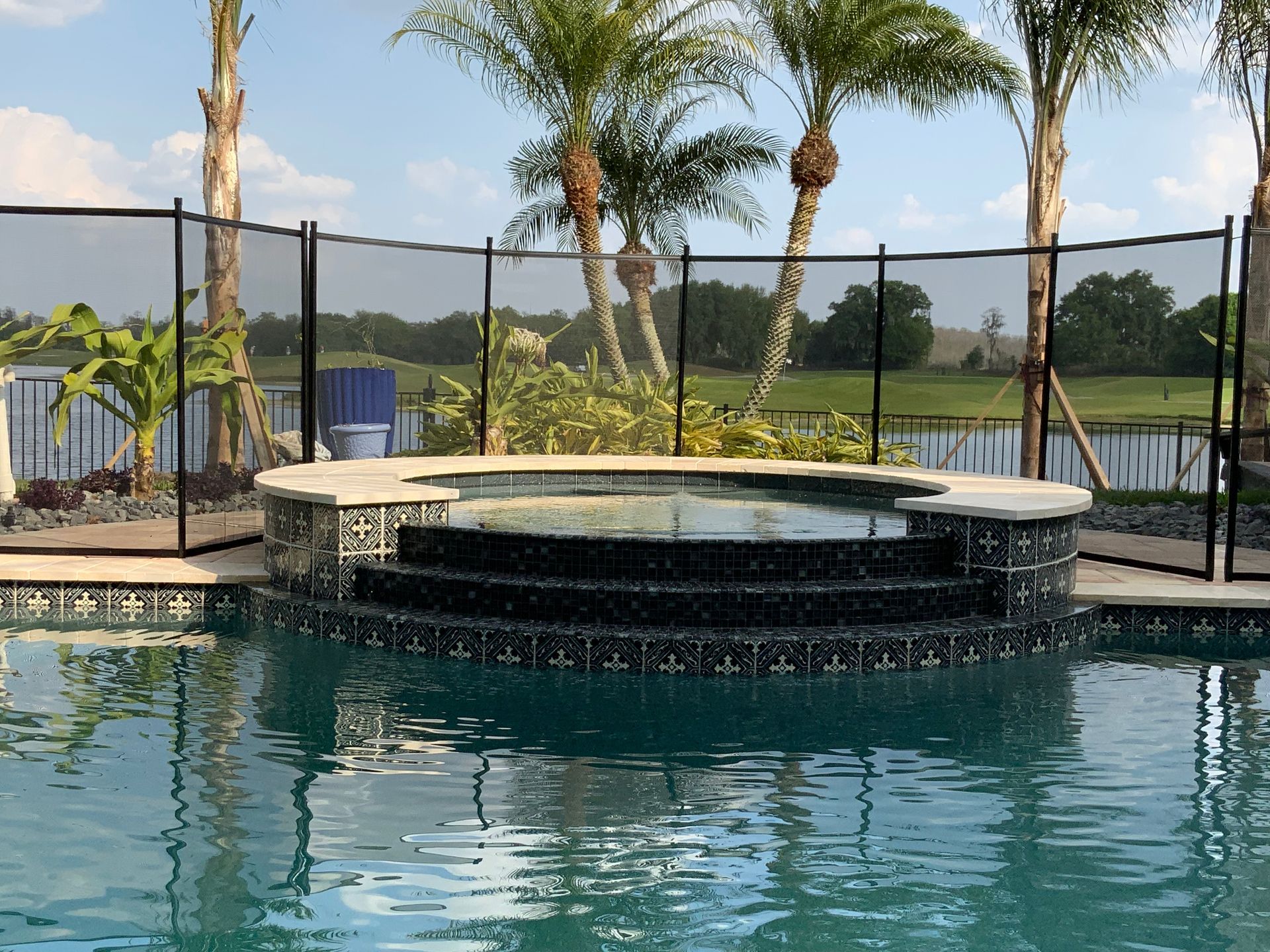 Swimming pool with a raised hot tub and safety fence. Palm trees and a lake are in the background.