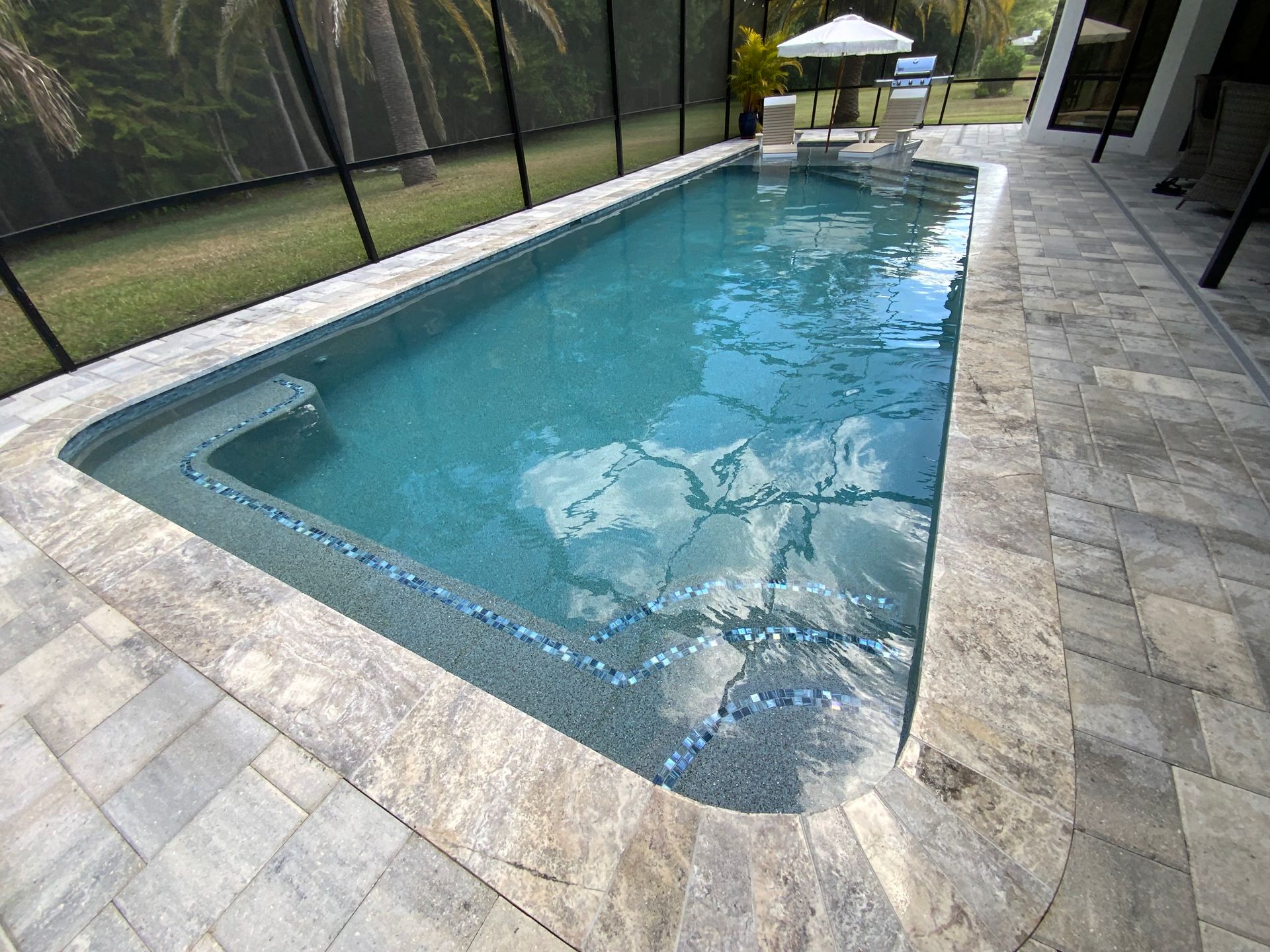 Rectangular swimming pool with light blue water surrounded by stone pavers, screened enclosure, and landscaping.