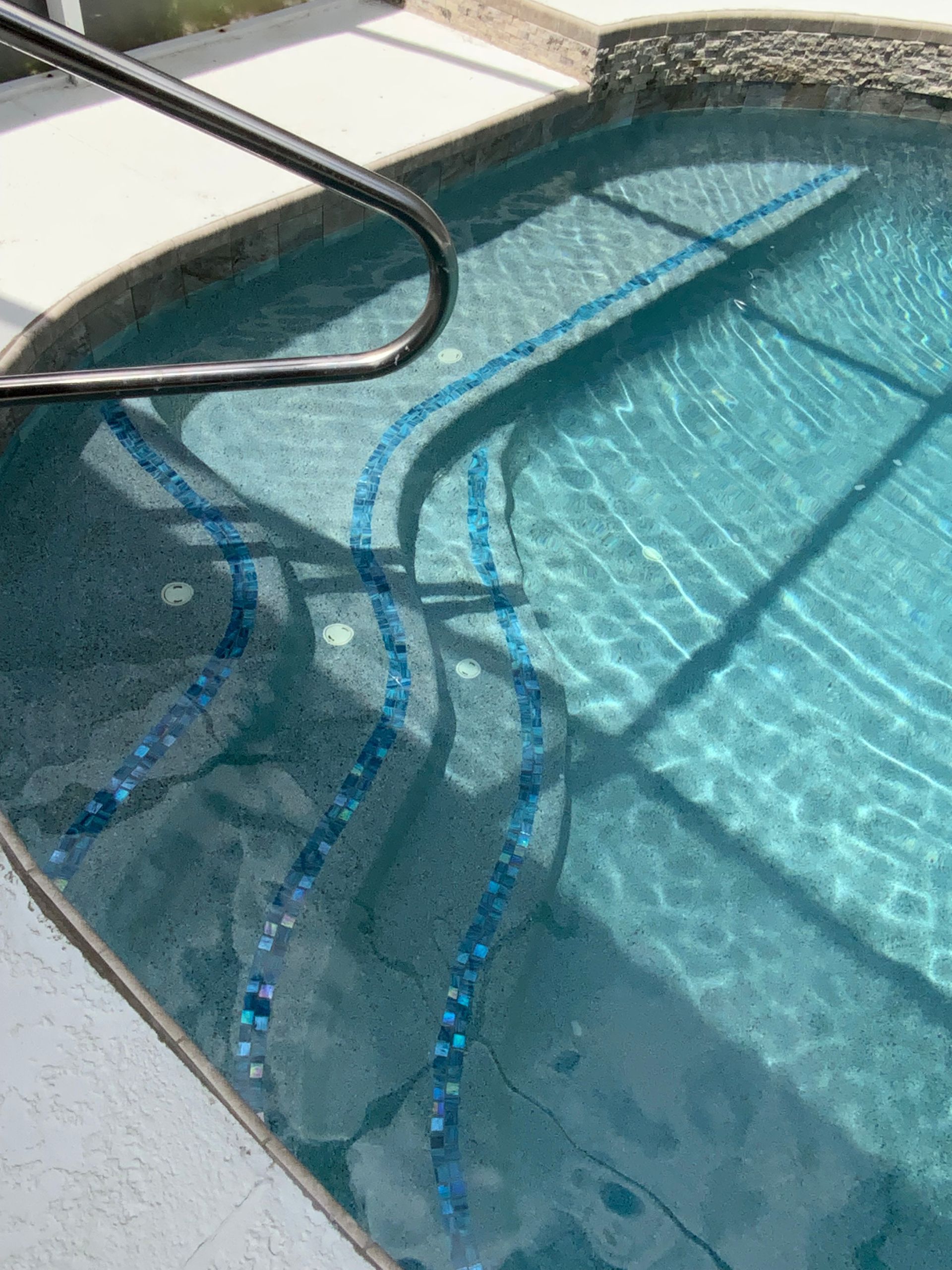 Pool with steps, blue tile accents, metal handrail. Water is clear, reflecting sunlight.