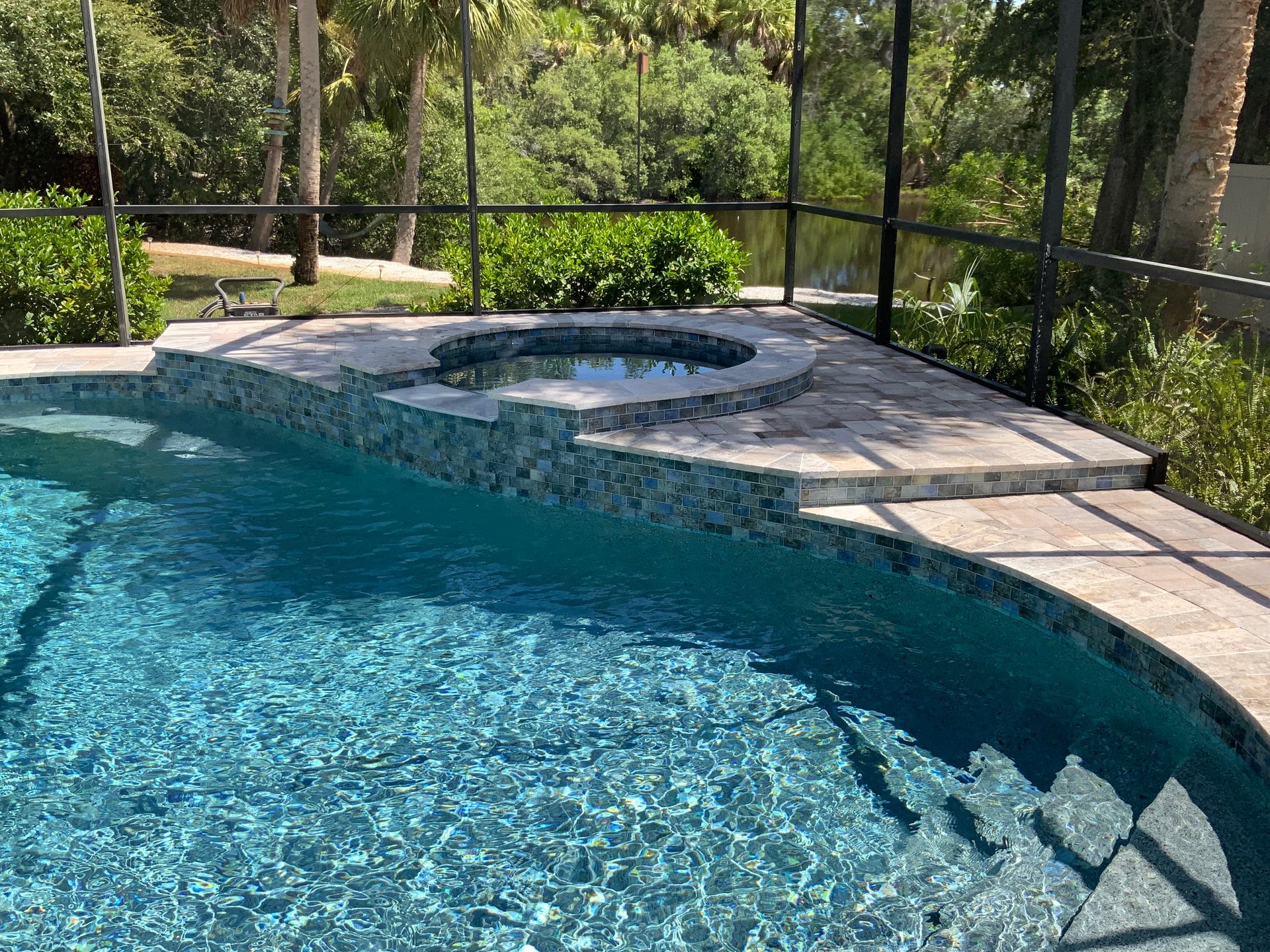 Pool with hot tub, stone patio, and lush green background.