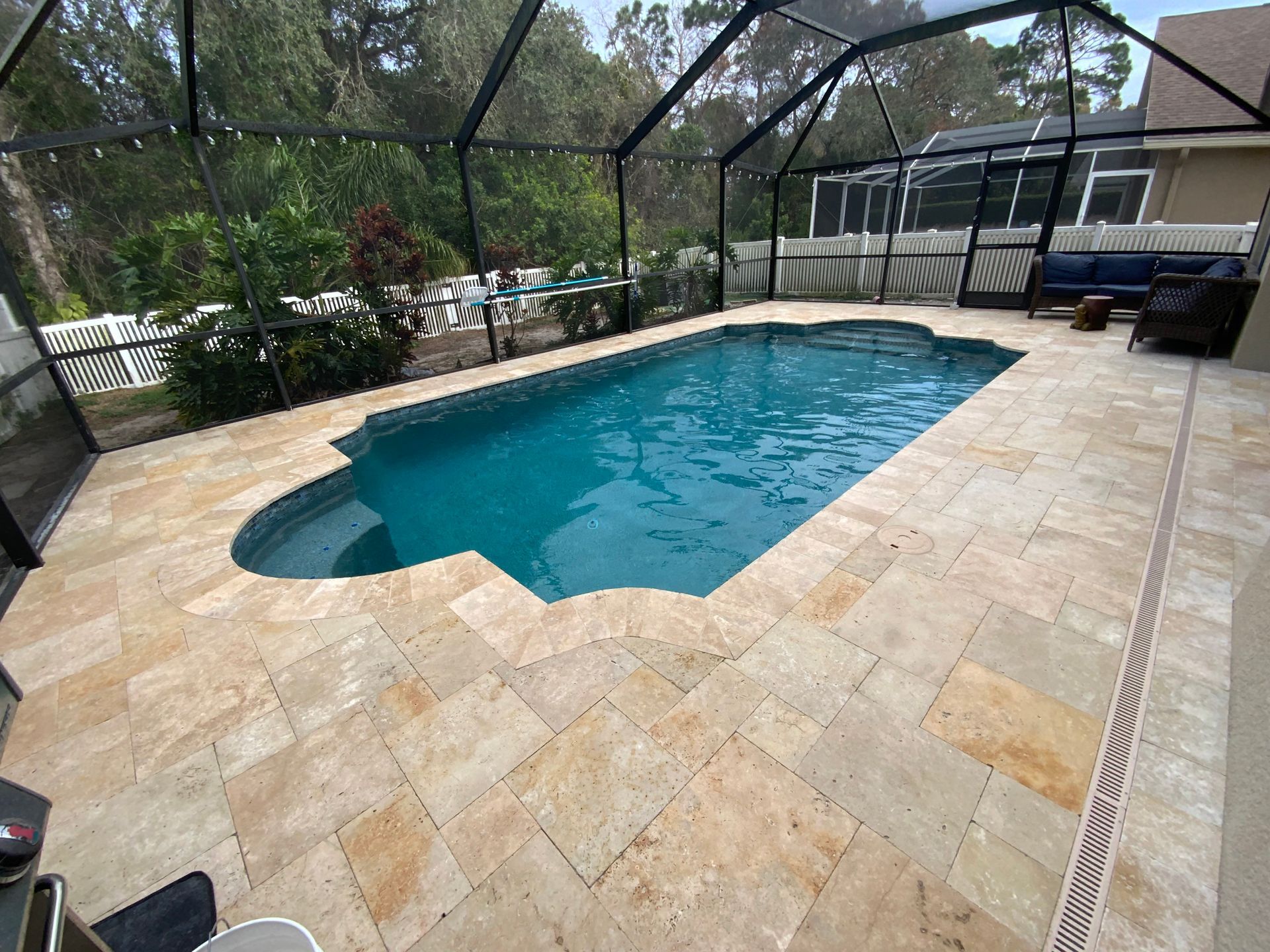 Pool with light-colored stone surround enclosed in a screen, surrounded by greenery.