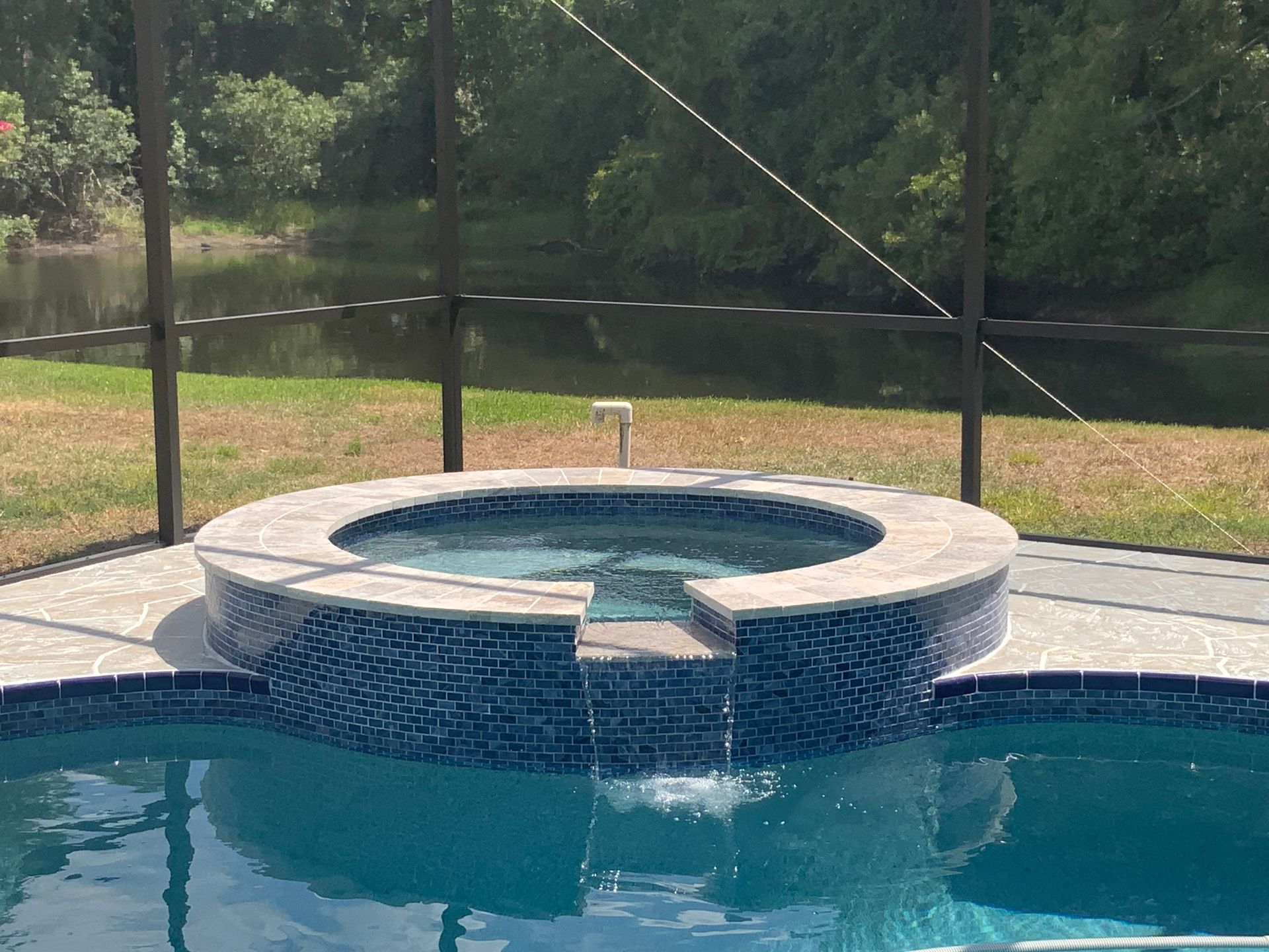 A pool with a blue-tiled hot tub and a small waterfall, in a screened enclosure, overlooking a lake.