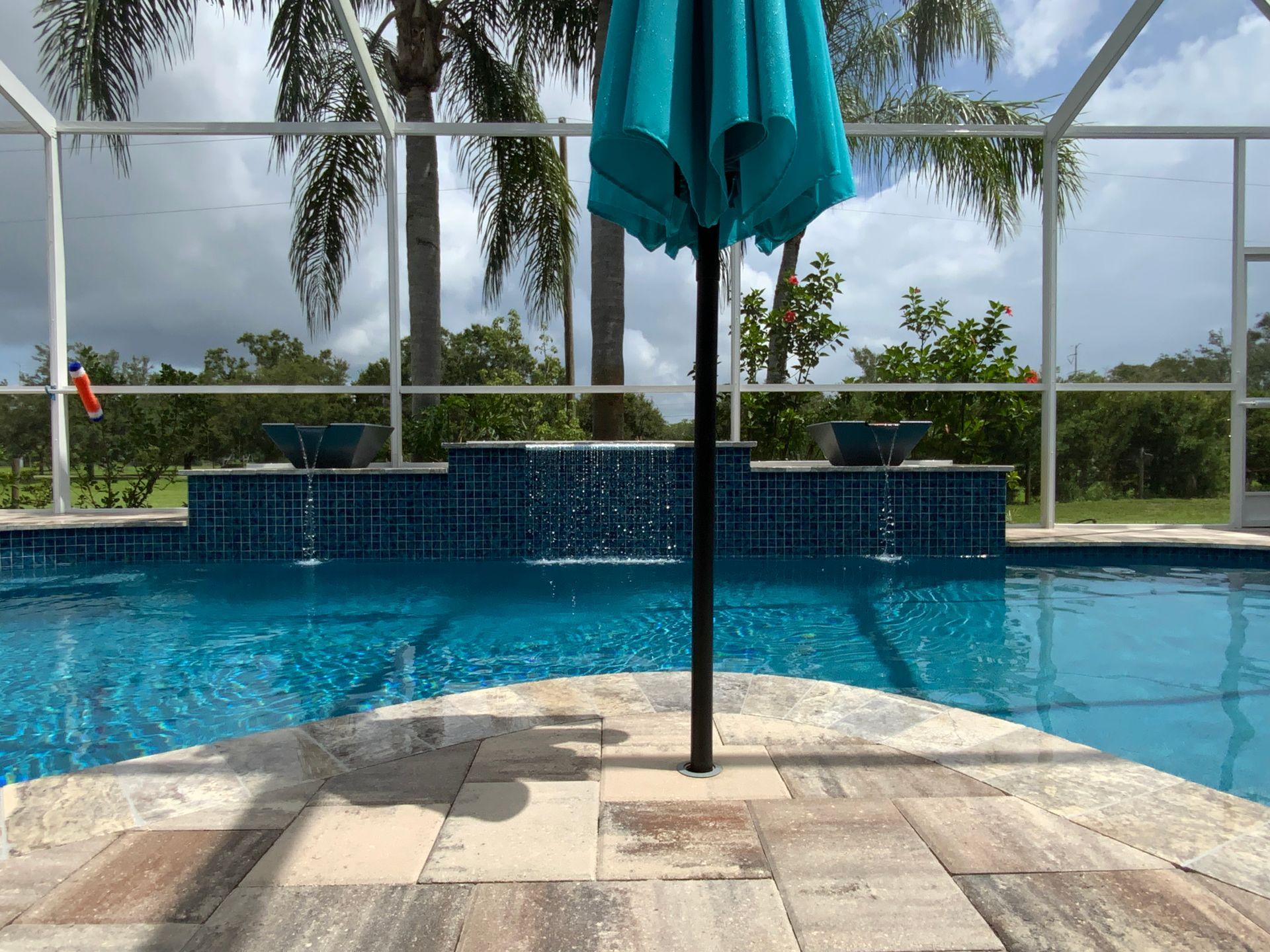 Pool with turquoise water, waterfall feature, patio, and teal umbrella, under a screened enclosure.