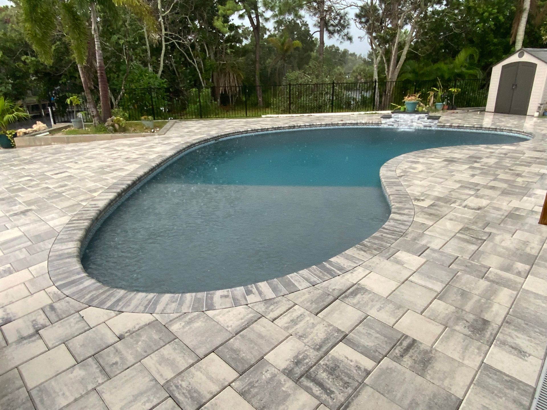 Pool with gray pavers and water feature, surrounded by trees and a small shed.
