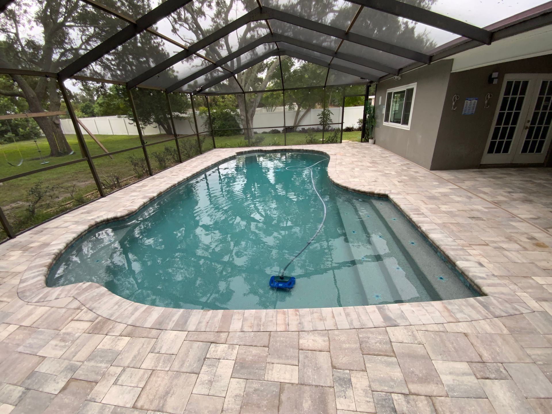 Screened-in pool with a curved design, surrounded by stone pavers, next to a home with double doors.