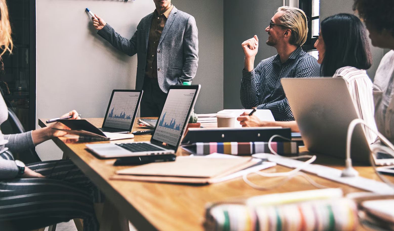 Business meeting; man presenting at whiteboard, colleagues with laptops at conference table.
