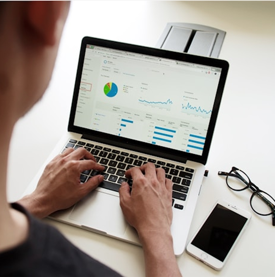 Person working on a laptop, analyzing data charts with a smartphone and glasses nearby.
