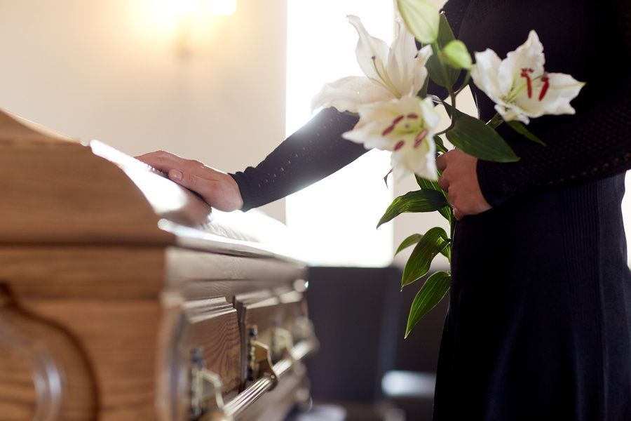 Person placing a hand on a wooden casket beside white lilies during a funeral