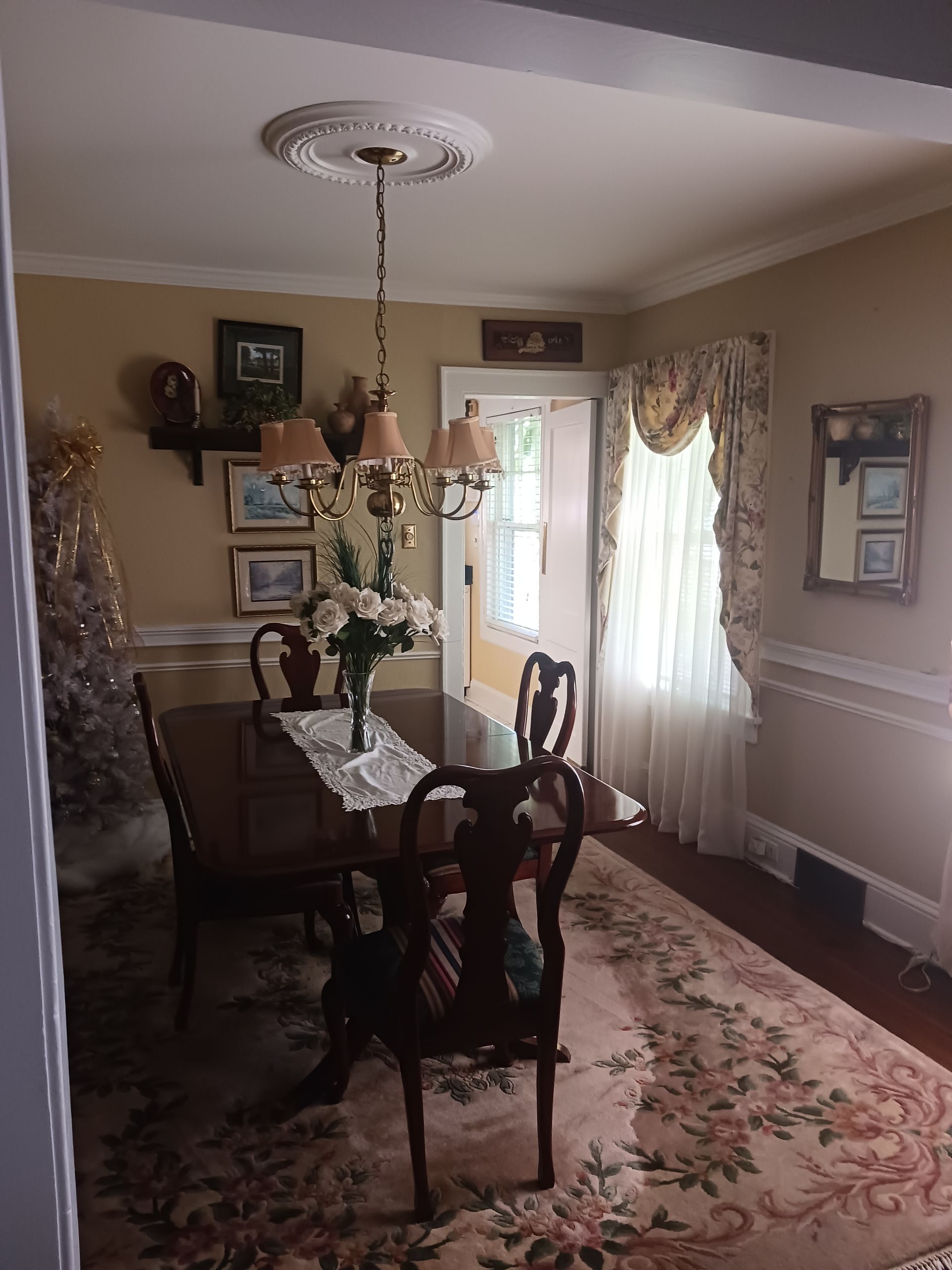 Dining room with a wooden table, ornate chairs, floral rug, and a chandelier. Pale yellow walls and a window with a floral curtain.