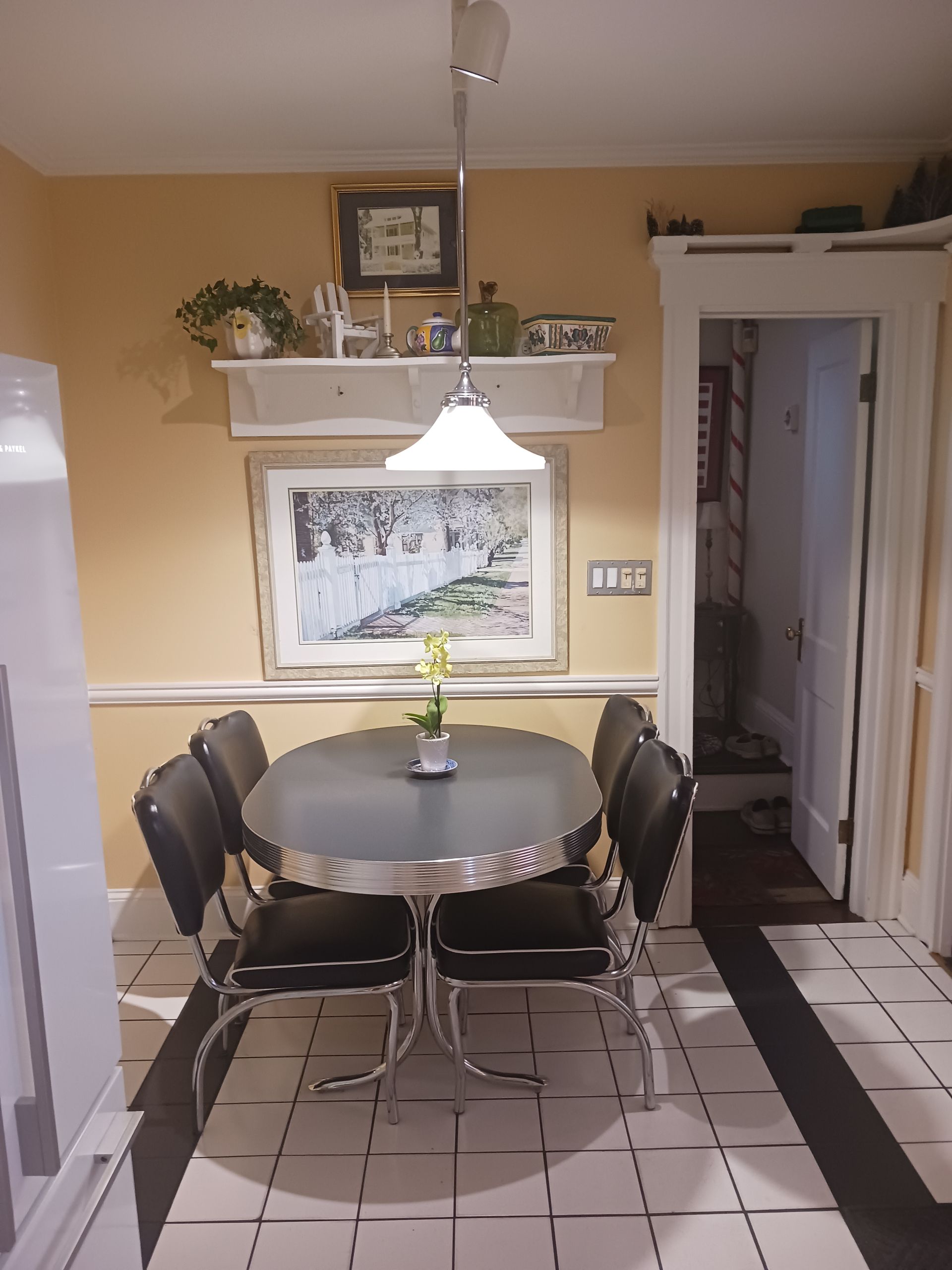 Dining area with an oval table and four black chairs, beneath a hanging light. Yellow walls, a framed picture, and open doorway visible.