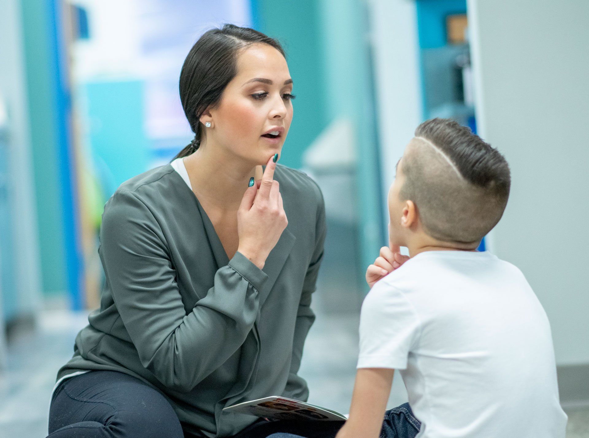 A young female Speech Therapist sits on the floor with her hand on her chin as she helps her young patient with his speech. She is dressed in semi-casual clothing and working with the young boy using a story book that is open in her hand. The boy is dressed casually and seated cross-legged on the floor as he looks up at the therapist and copies her words with his hand on his own chin.