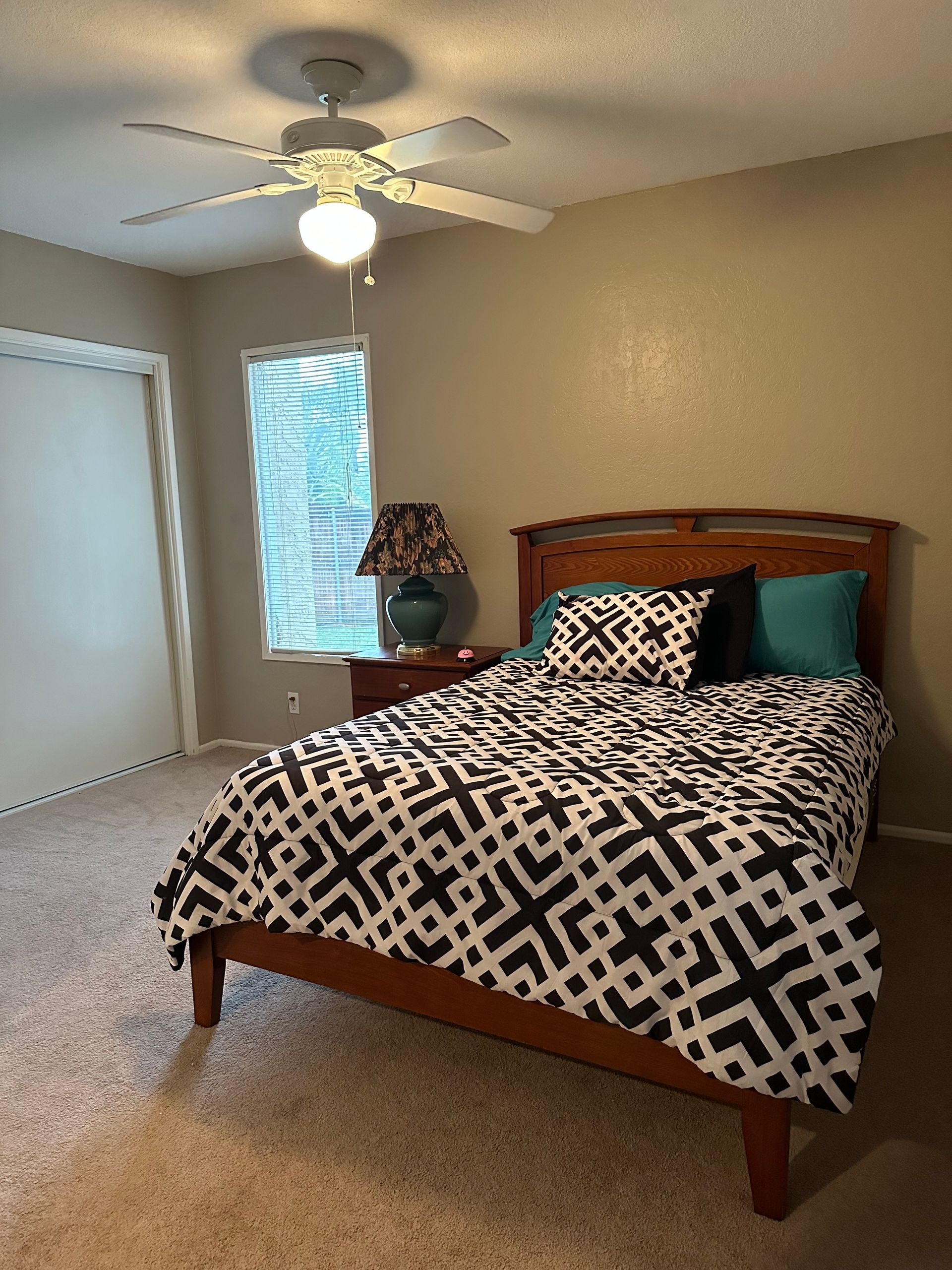 Bedroom with bed, patterned bedding, nightstand with lamp, window, closet, ceiling fan.