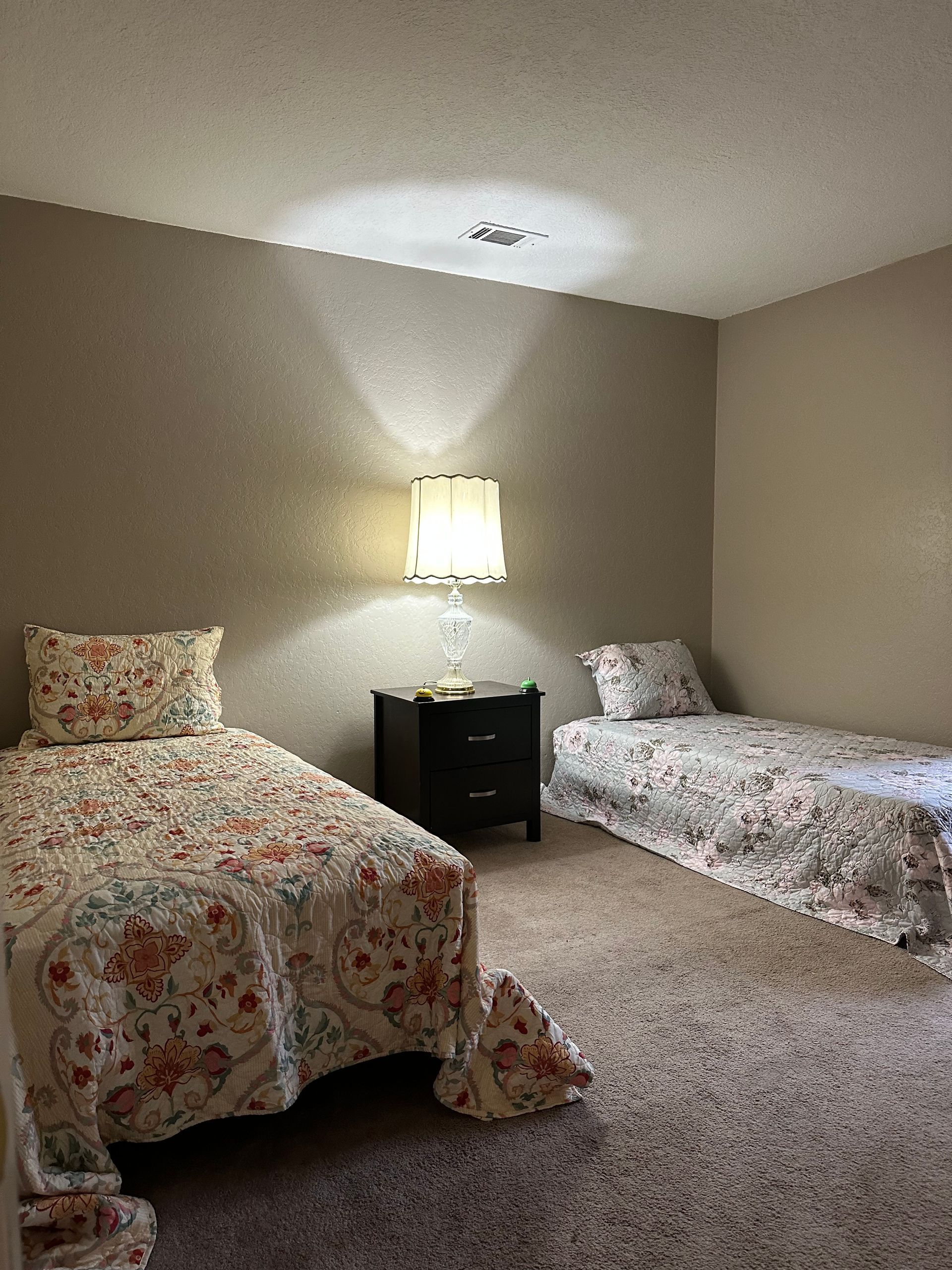 Two twin beds in a room with a nightstand and lamp between them. Floral bedspreads and a neutral-toned wall.